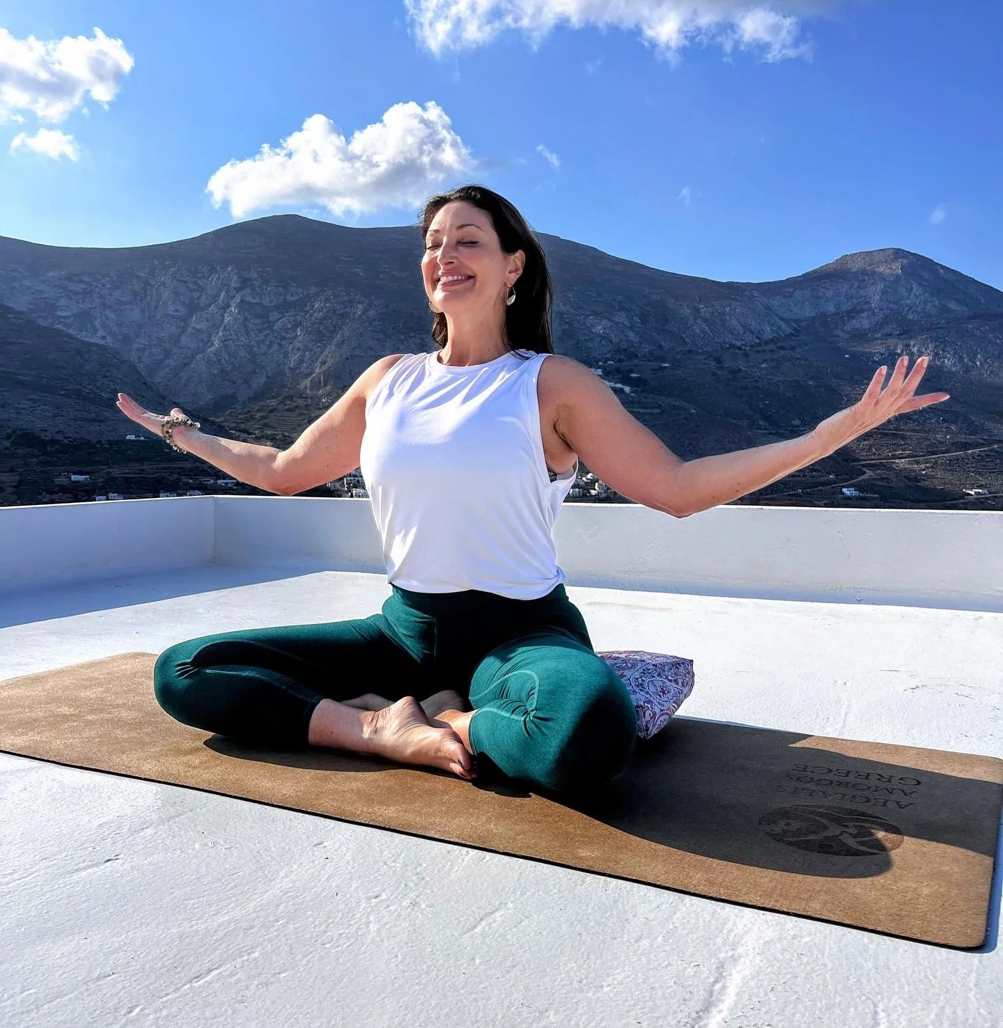Woman practicing yoga on rooftop with mountain view, sitting cross-legged on mat, arms outstretched, smiling, sunny day