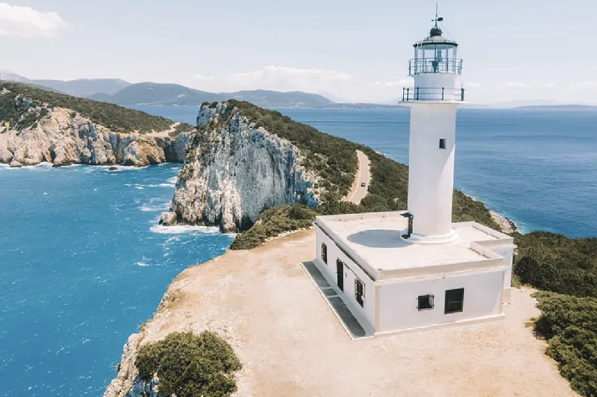 White lighthouse on a cliff overlooking the ocean with rocky formations and a distant shoreline.
