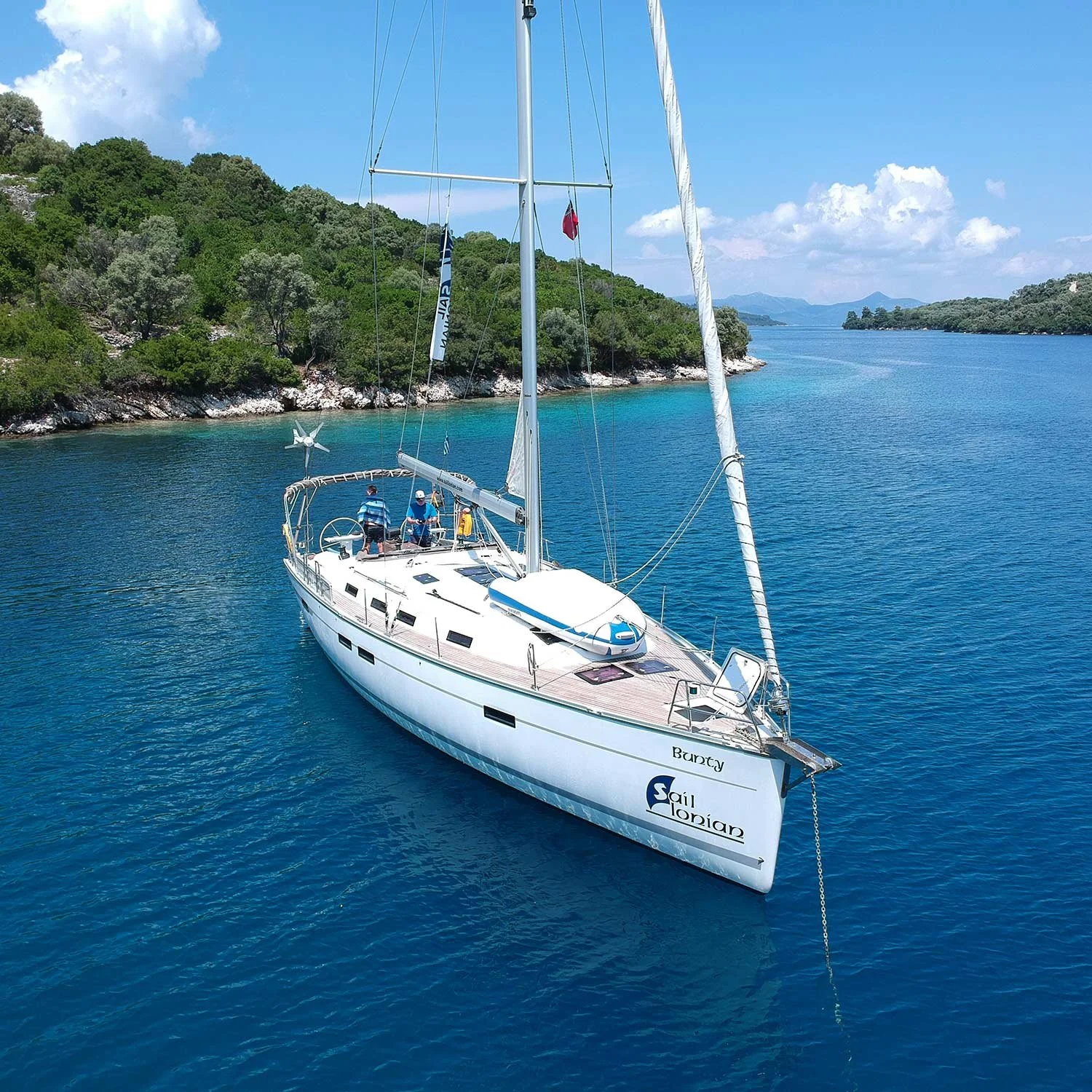A white sailboat named 'Bounty' sailing in calm blue waters near a green, forested coastline under a partly cloudy sky.