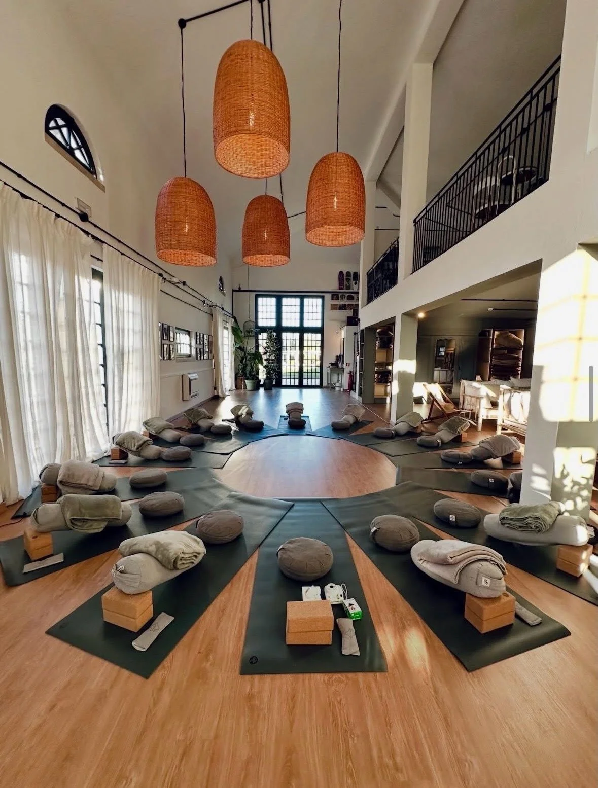 A spacious yoga studio with mats, bolsters, and blankets arranged in a circle on a wooden floor, illuminated by natural light and four large wicker pendant lights.
