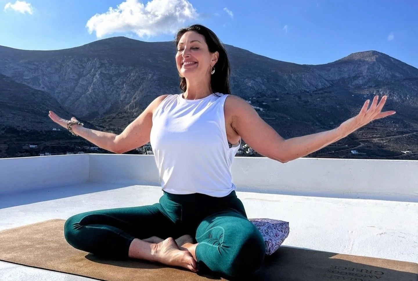 Woman meditating outdoors on a yoga mat with mountain landscape in the background, wearing a white sleeveless top and green leggings, smiling with arms outstretched.