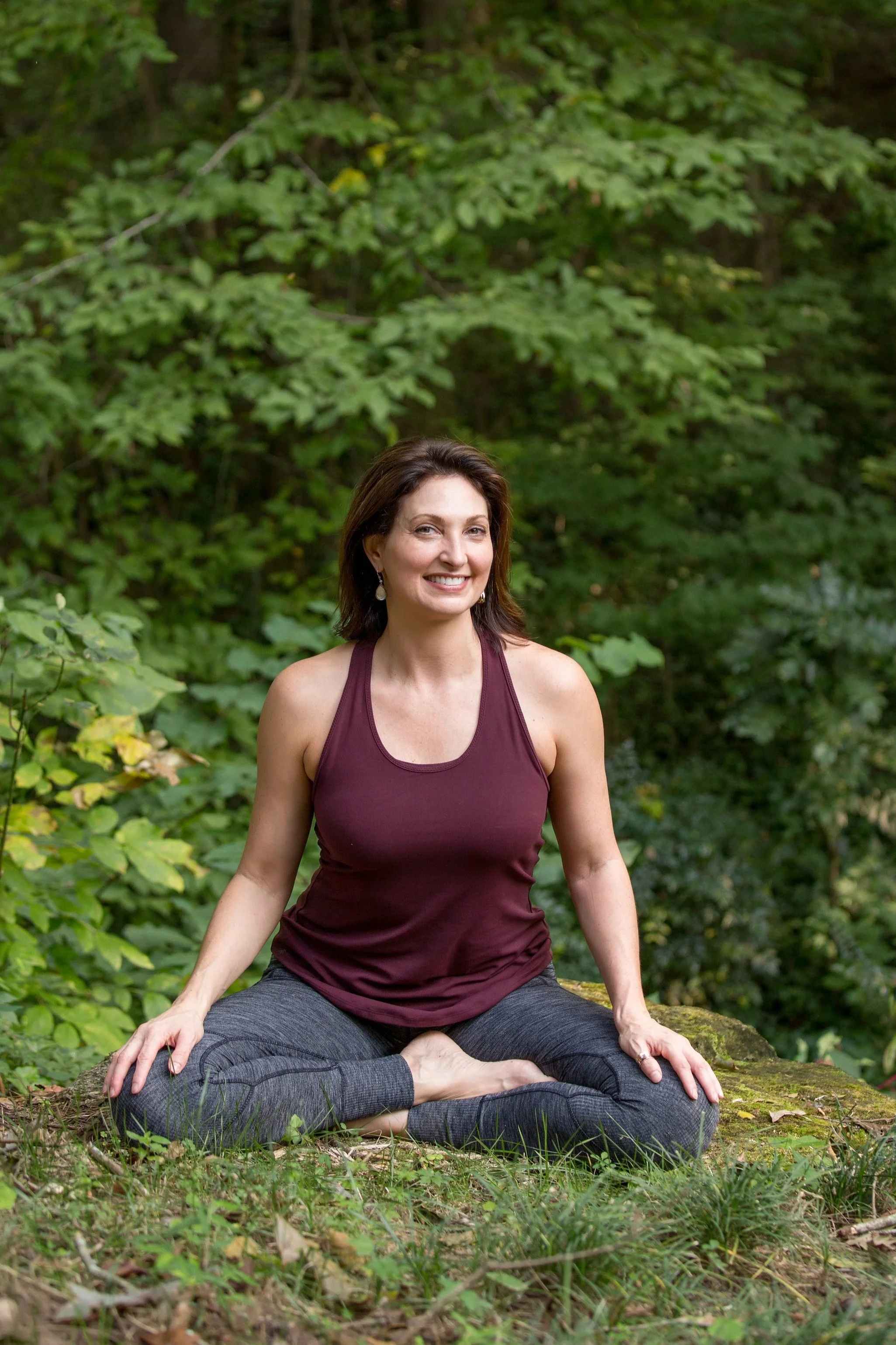 Woman practicing yoga outdoors on a moss-covered rock, sitting in a cross-legged meditation pose surrounded by green trees and foliage.