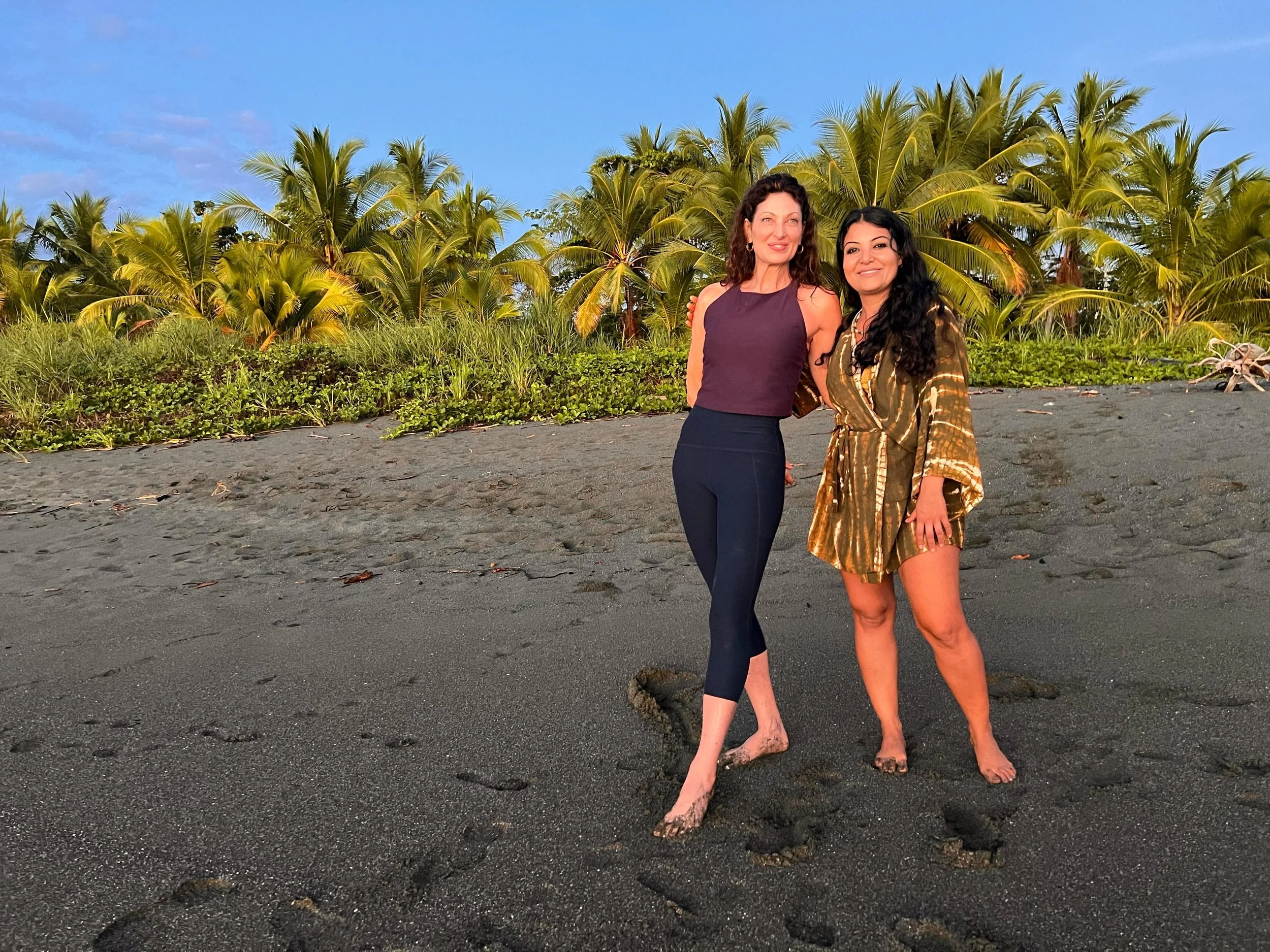 Two women standing together on a sandy beach with palm trees in the background, smiling at the camera, both barefoot and with dark hair, one wearing a sleeveless top and the other a patterned dress.