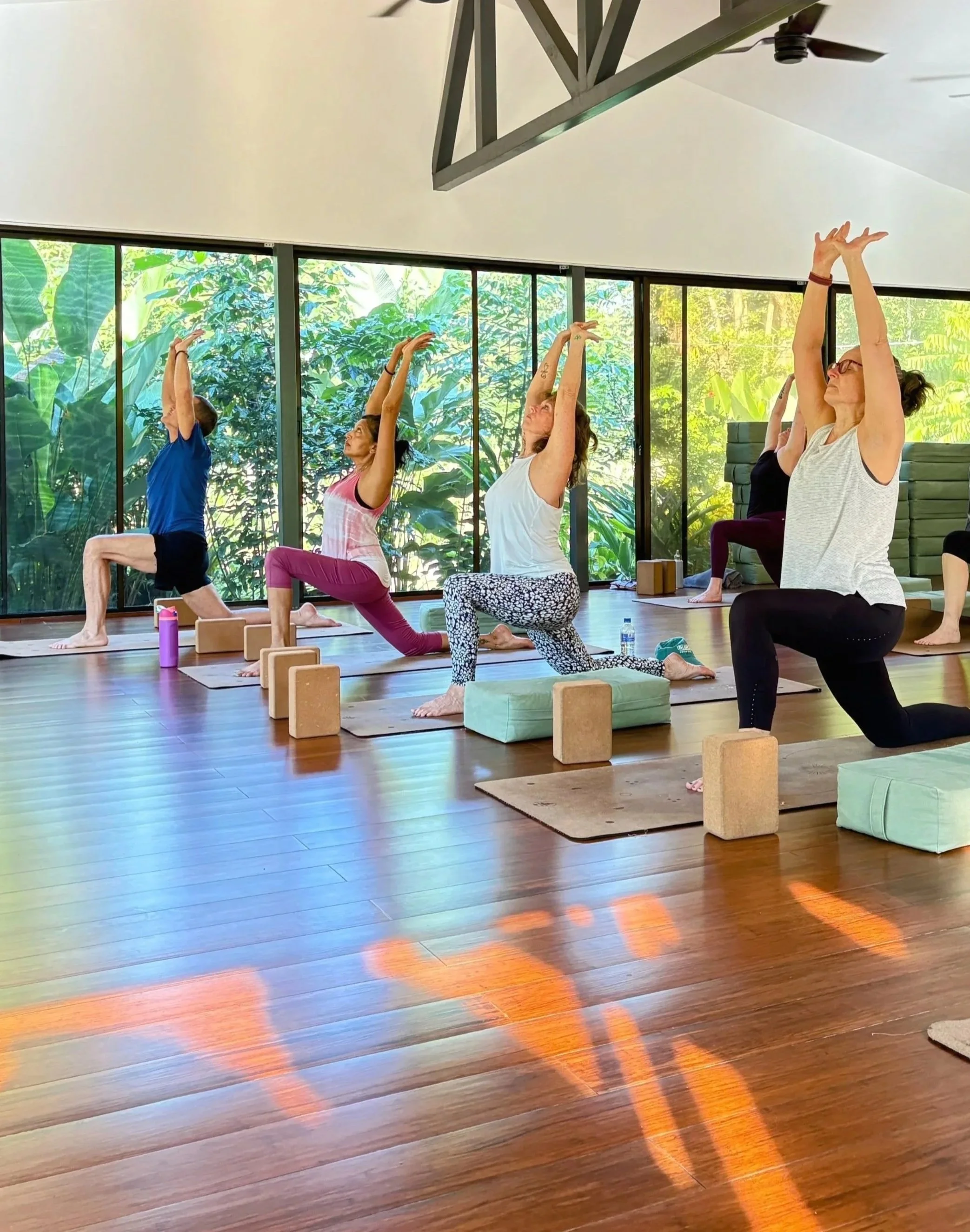 Four women practicing yoga on mats in a room with large windows overlooking a green outdoor area.