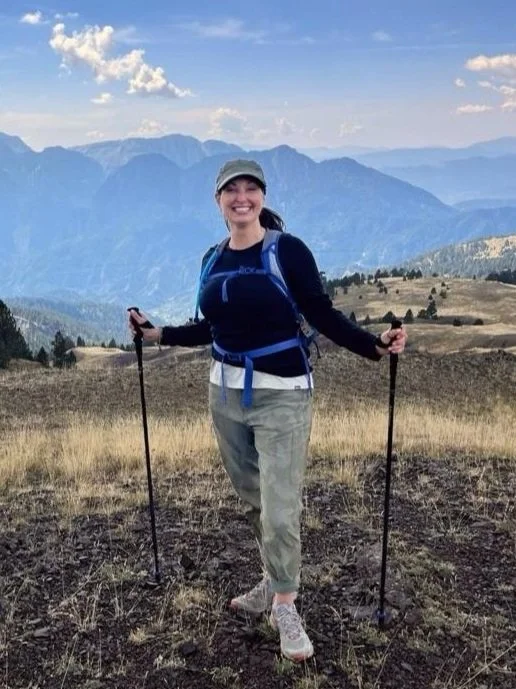 Woman hiking in mountains smiling, holding trekking poles, wearing a cap, backpack, and outdoor clothing, with mountain range in the background.