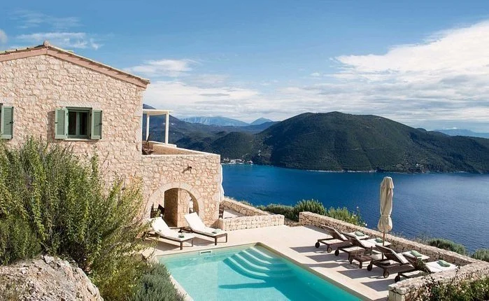A stone house with green shutters next to a swimming pool, overlooking a lake and mountainous landscape under a partly cloudy sky.