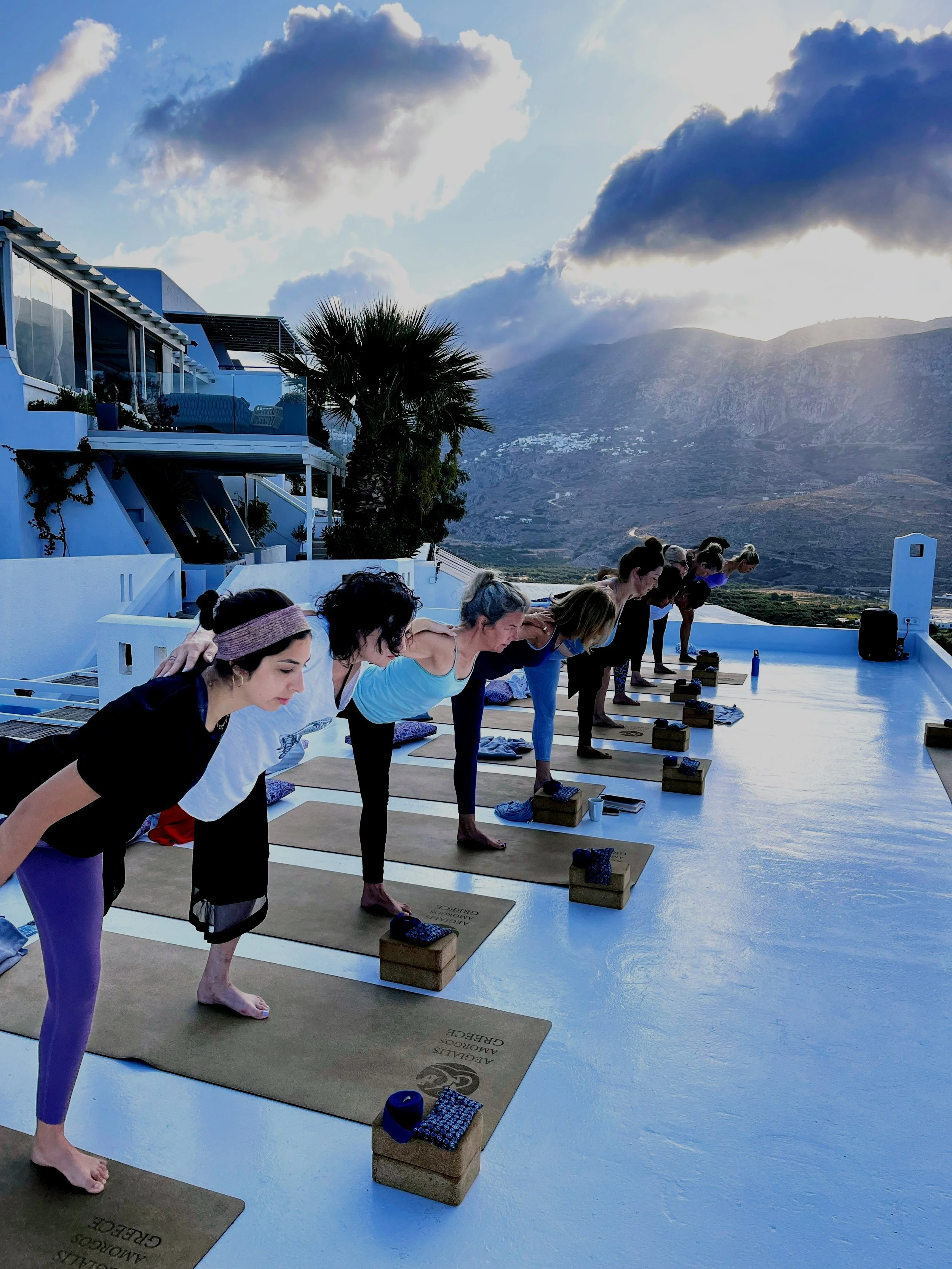 A group of women practicing yoga on mats on a rooftop terrace with a scenic mountain view and cloudy sky.