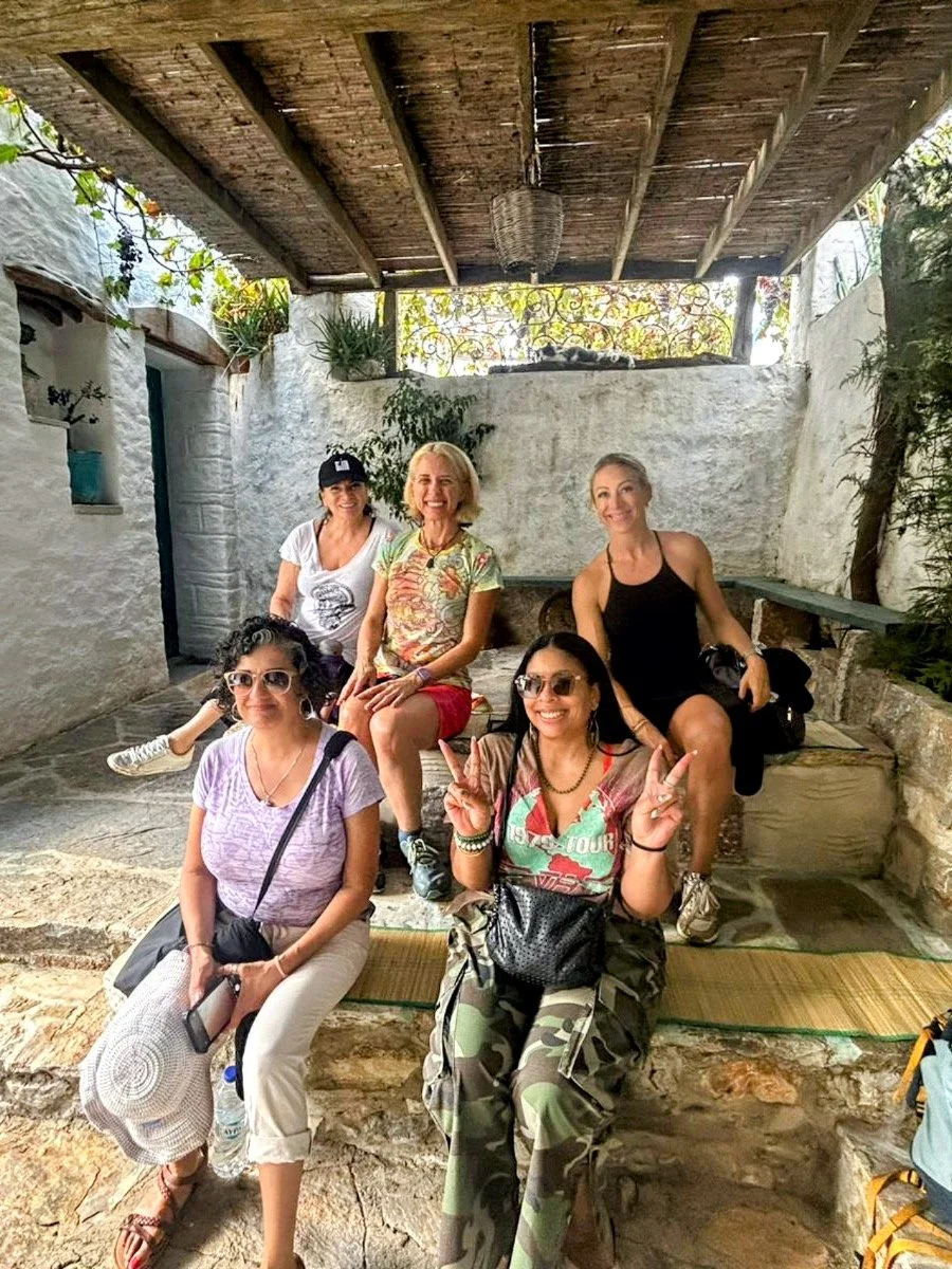 Five women sitting and posing for a photo in an outdoor setting with rustic white walls and a wooden roof. The women are smiling, with some making peace signs, and there are various plants around them.