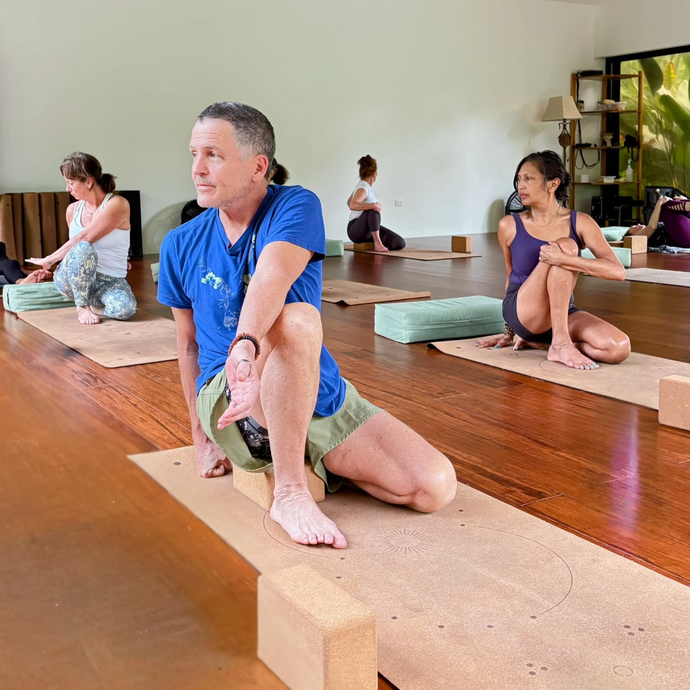 Two people practicing yoga indoors on mats, one kneeling with hands in a prayer position and the other in a seated pose with hands extending forward, both in a serene room with wooden floors.