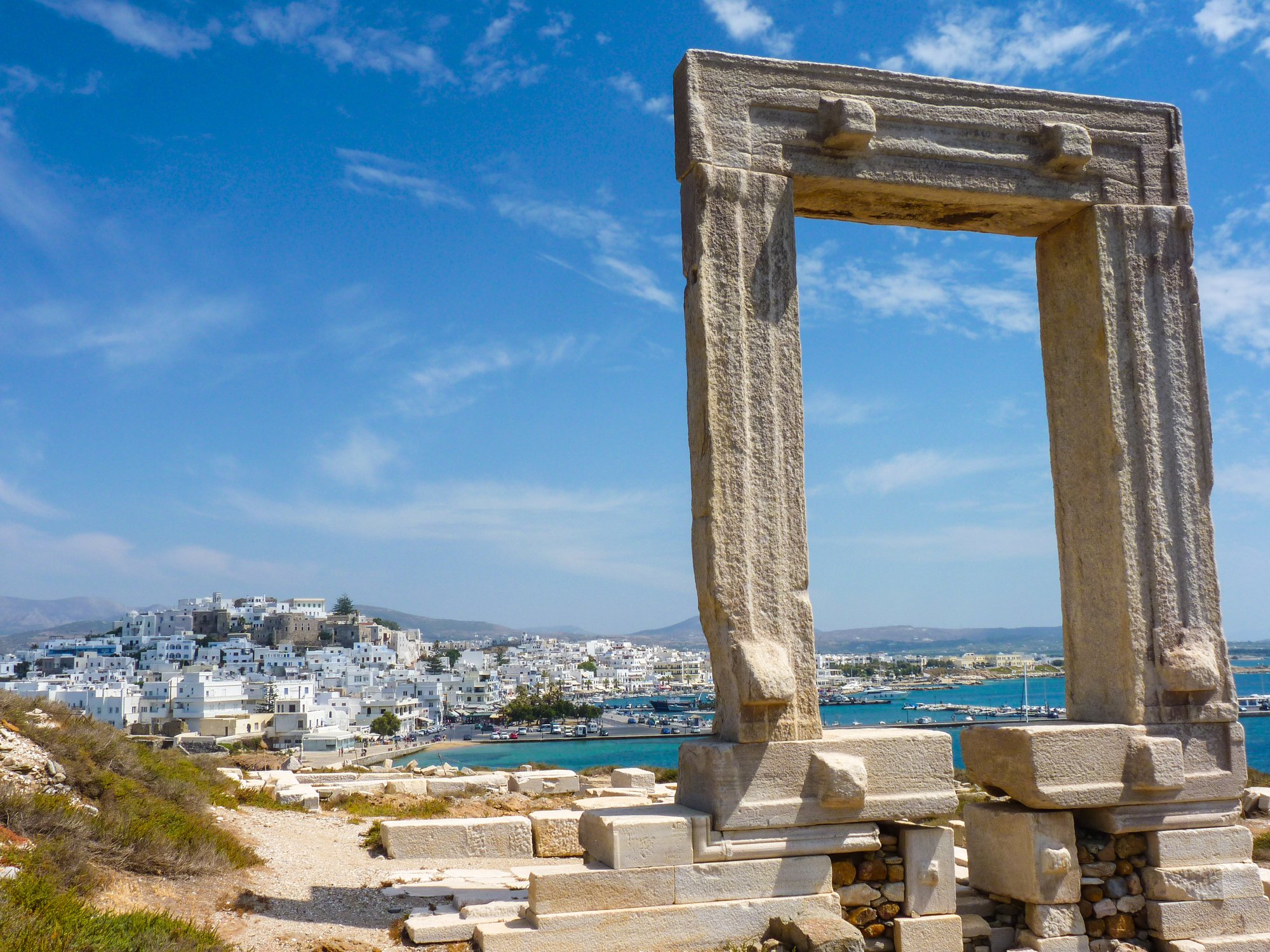 Ancient stone gateway overlooking a coastal town with white buildings, blue sky, and harbor.