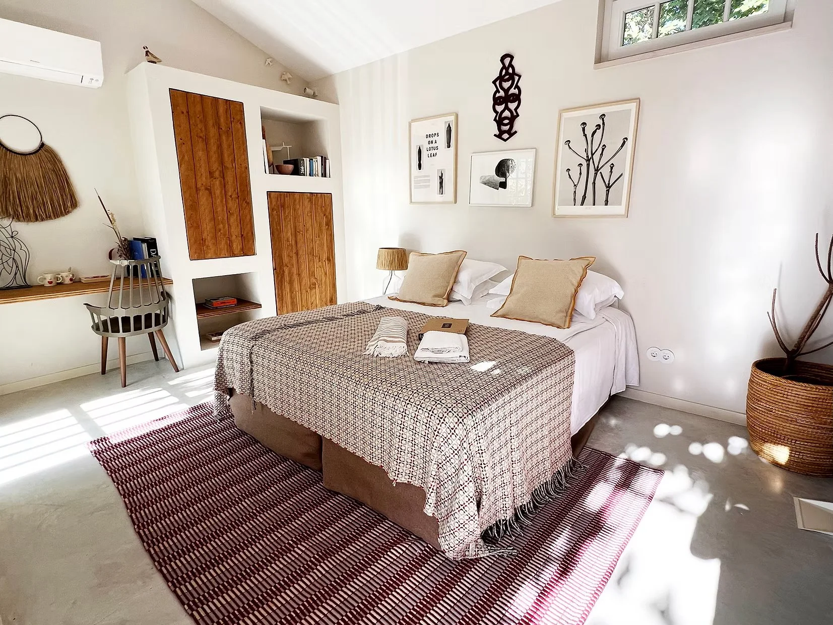 Bright bedroom with a bed covered in a patterned beige and red tablecloth, four pillows, and a red and white rug. A small side table with a lamp, framed artwork on the wall, and a large woven basket with a plant are visible.