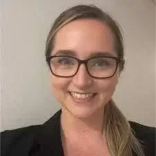 A woman with glasses smiling, with blonde hair tied back, wearing a black blazer, against a plain light-colored background.