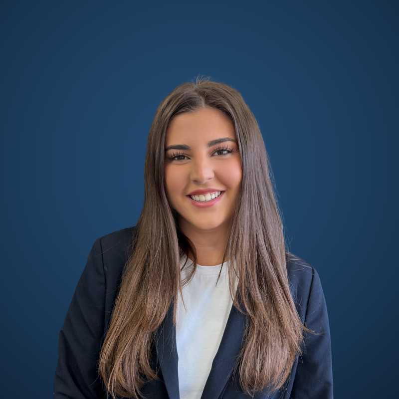 A woman with long brown hair smiling, wearing a white shirt and dark blazer against a plain dark blue background.