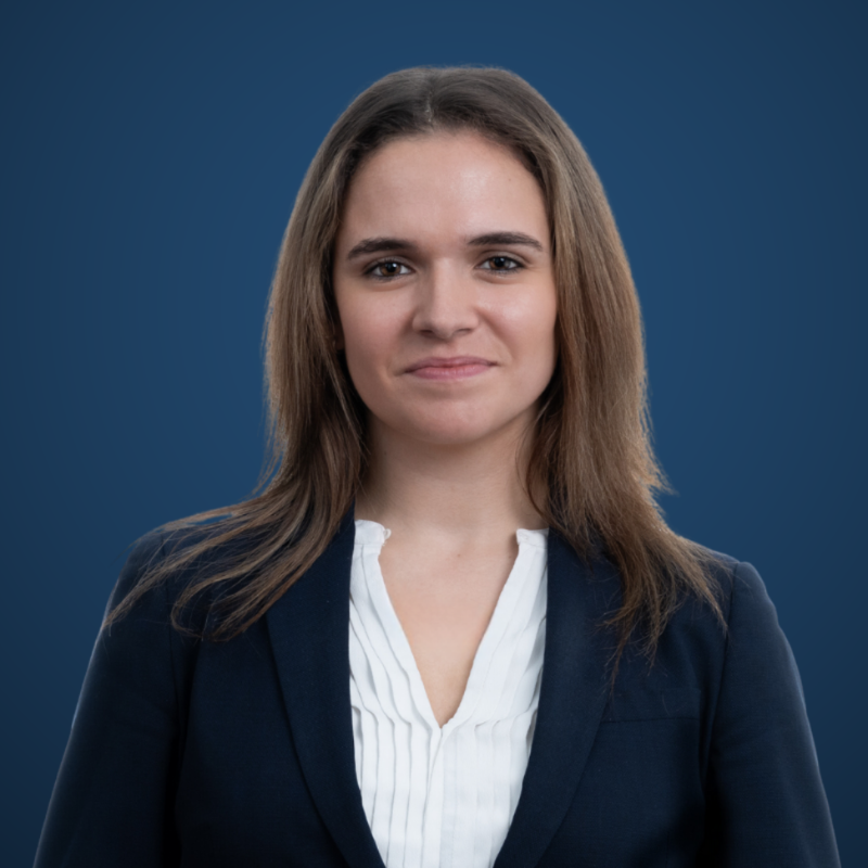 A portrait of a young woman with brown hair, wearing a navy blazer over a white blouse, against a blue background.