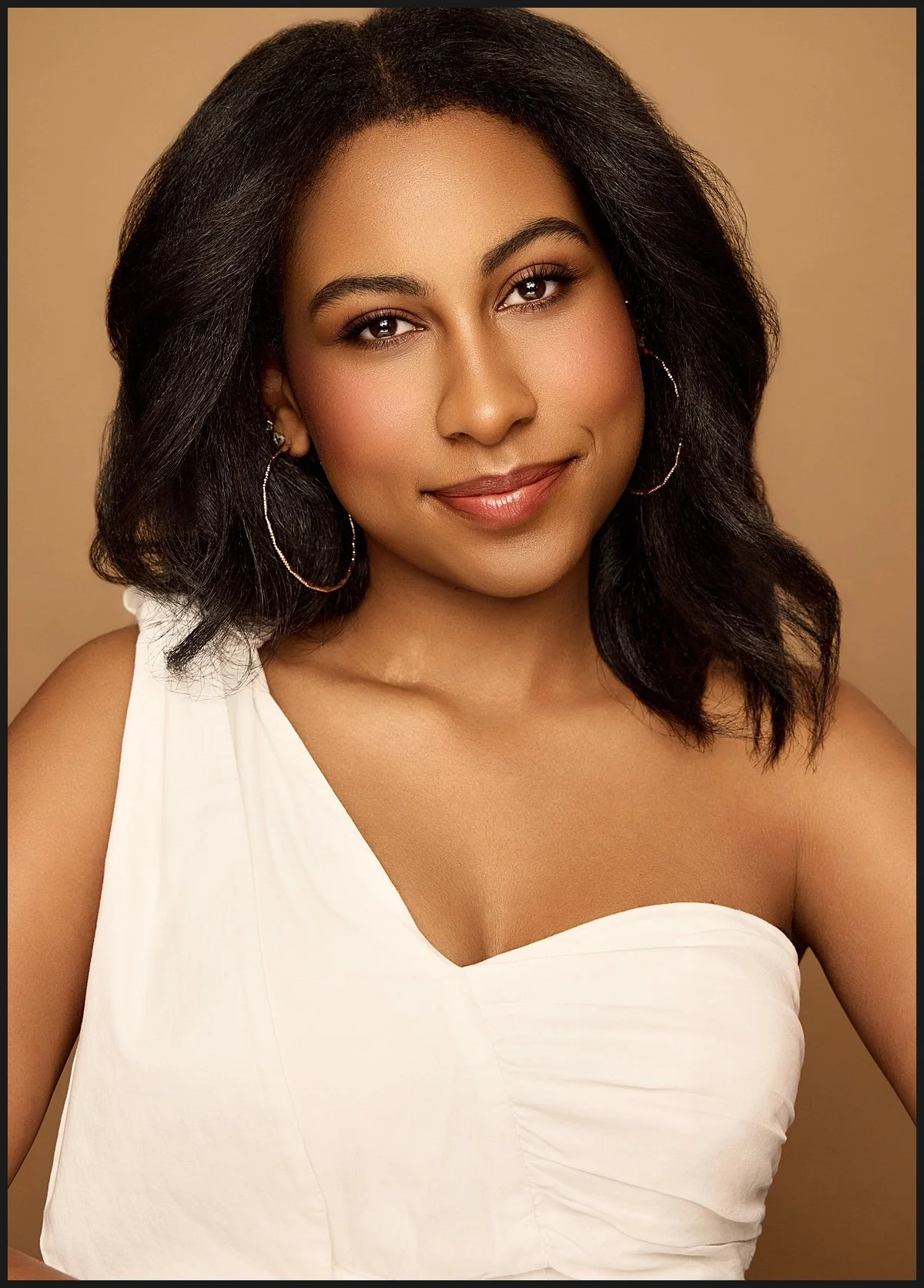 A woman with medium-length black hair, wearing hoop earrings and a white, one-shoulder top, smiling against a beige background.