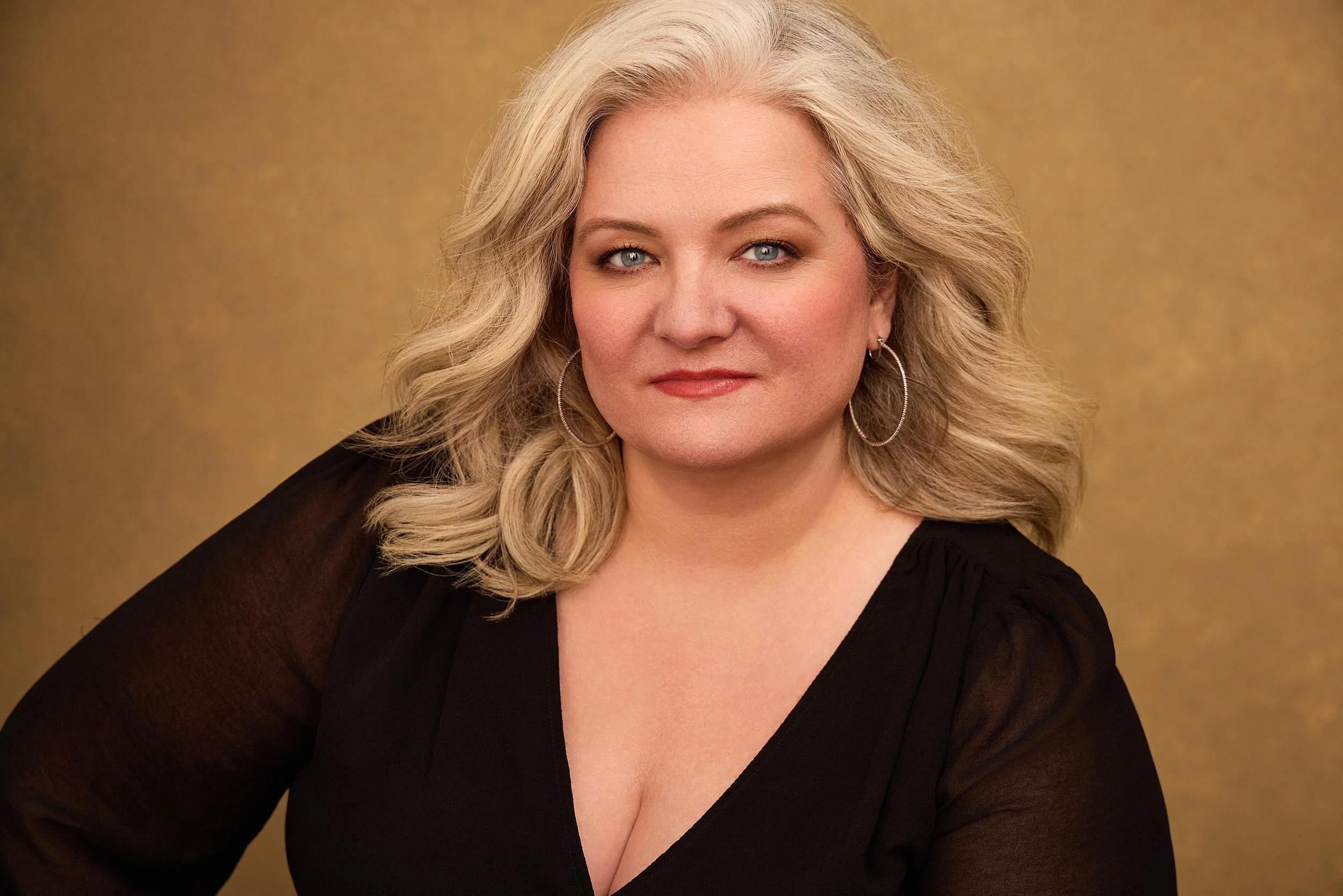 A woman with shoulder-length blonde hair, blue eyes, and hoop earrings, wearing a black top, posed against a gold-colored background.