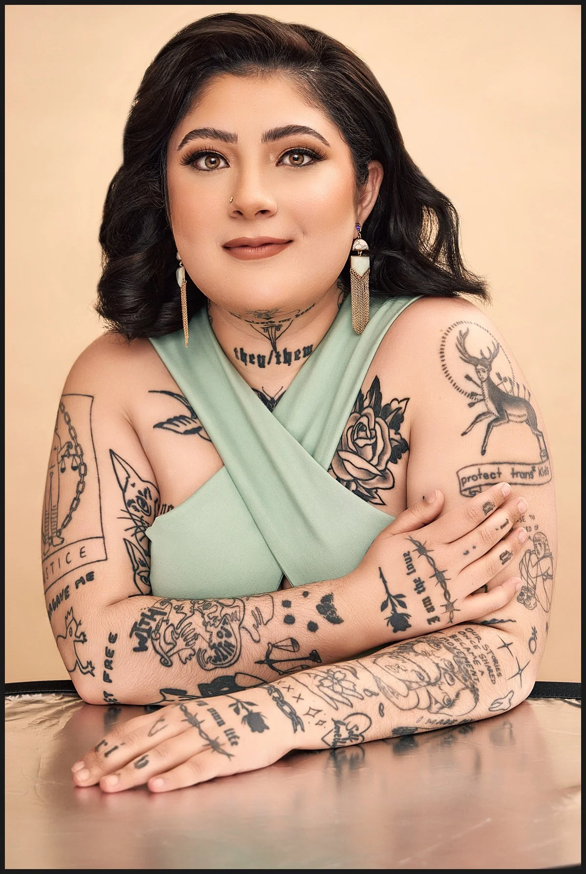 Portrait of a woman with dark hair, tattoos on both arms and chest, wearing a light green dress, and earrings, sitting at a reflective surface.