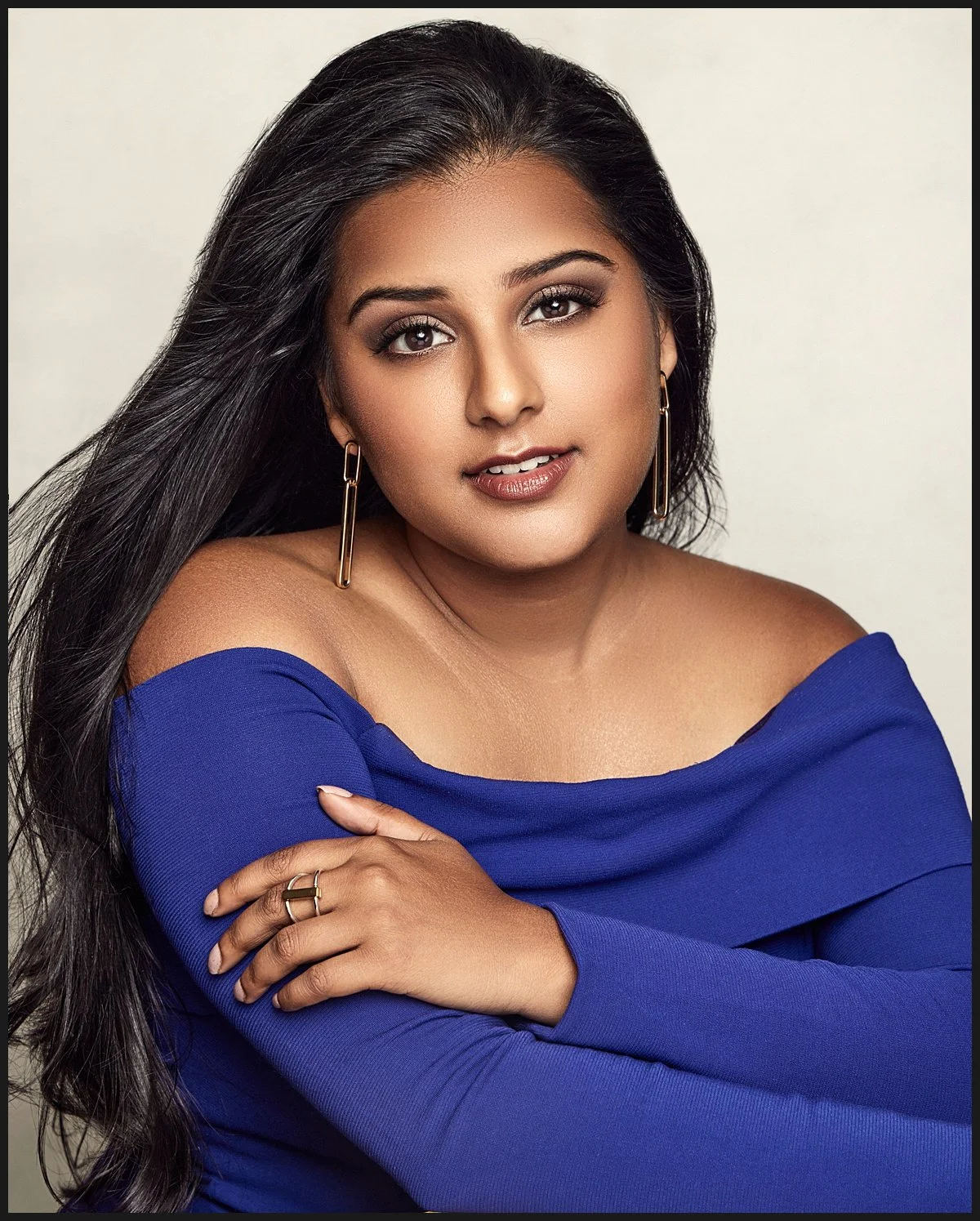 Portrait of a woman with long dark hair, wearing a royal blue off-the-shoulder top, gold hoop earrings, and a ring, against a light neutral background.
