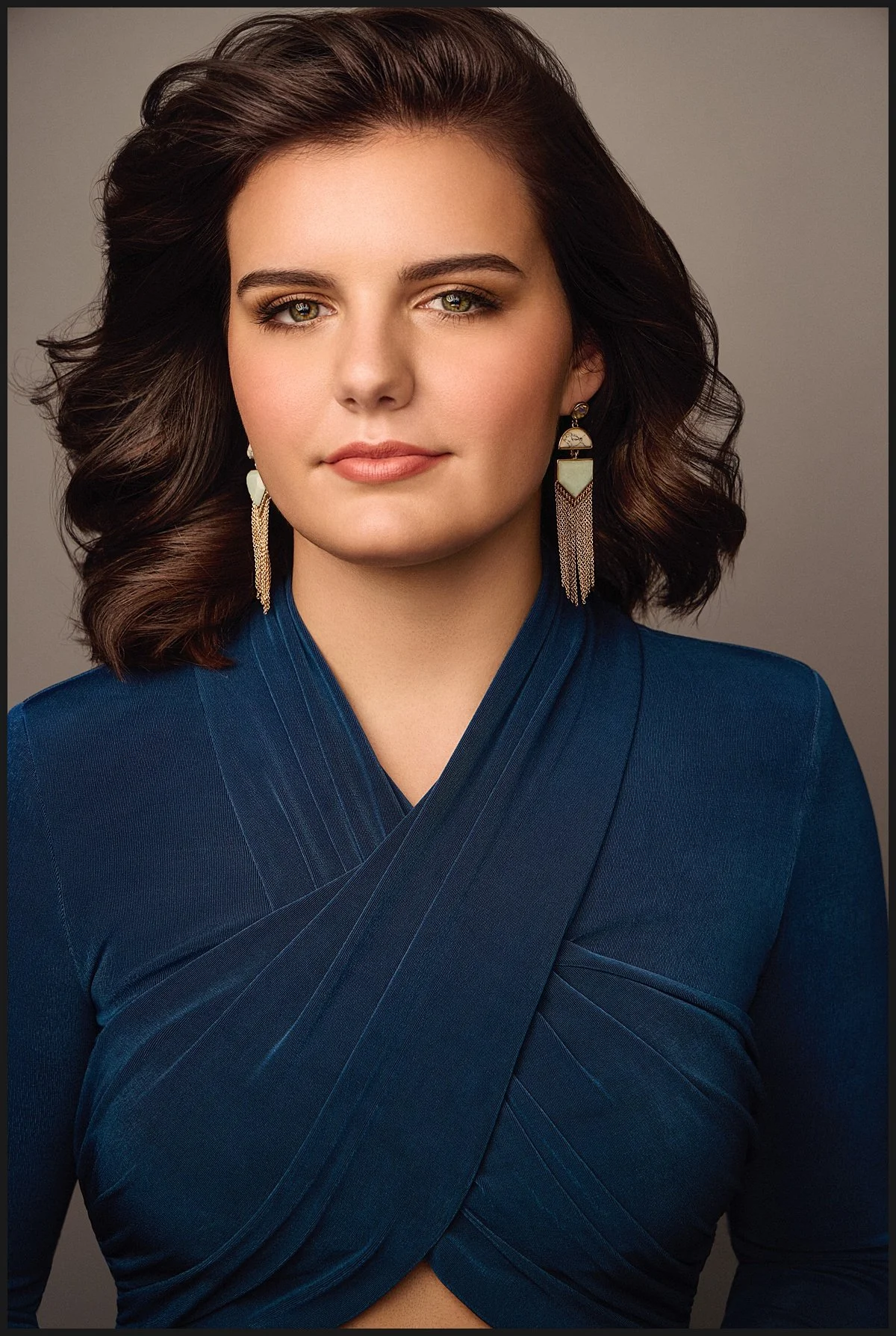 A woman with brown, wavy hair wearing a blue top with a wrap design and gold earrings.