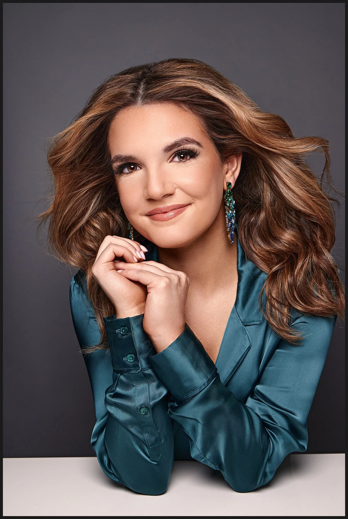 Portrait of a woman with wavy brown hair, wearing teal satin shirt, and large colorful earrings, smiling softly against a dark gray background.