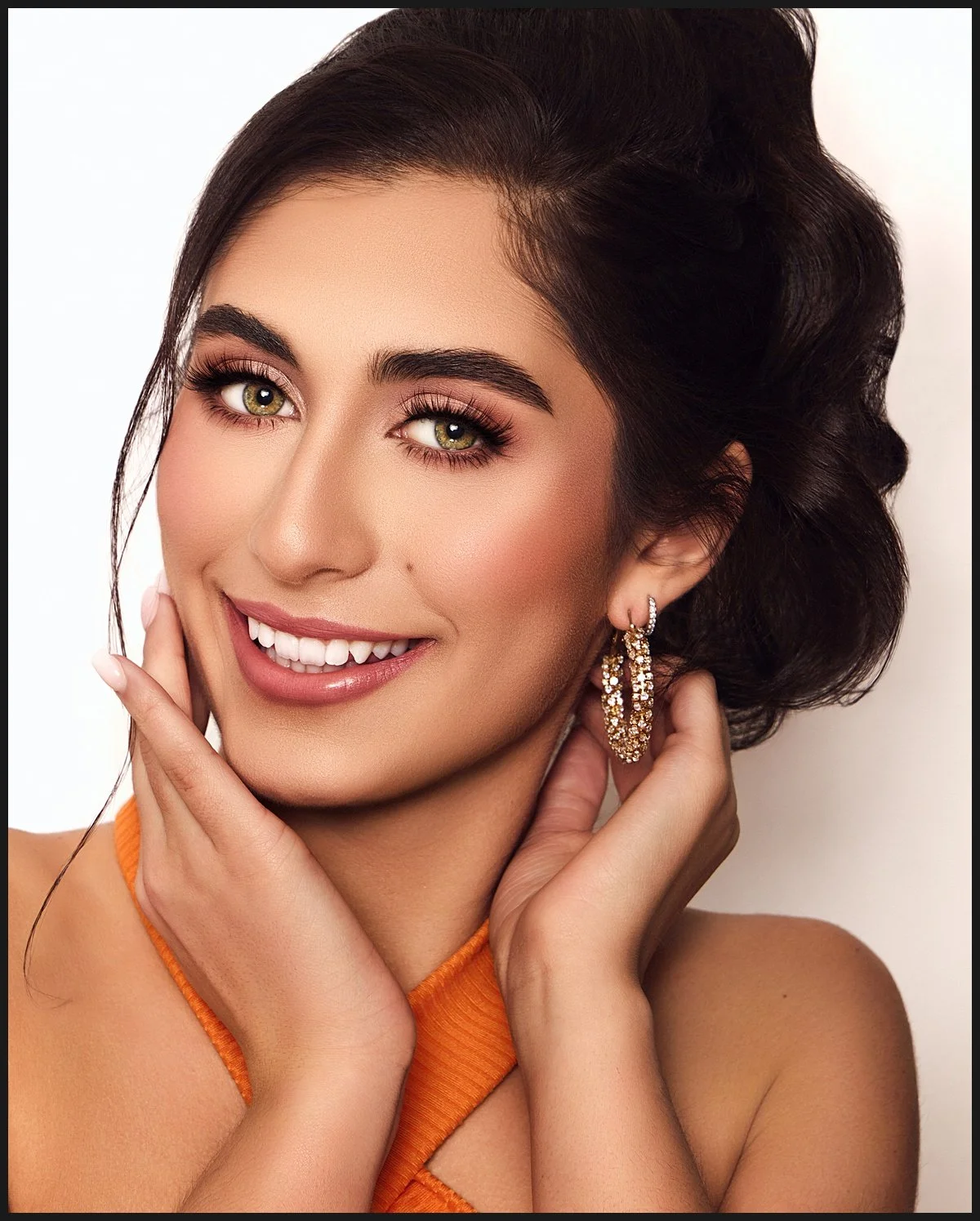 Close-up of a young woman with long dark hair, green eyes, and makeup, smiling, wearing gold earrings and an orange top.