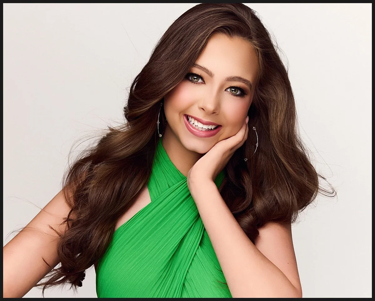 Portrait of a young woman with long wavy brown hair, smiling, wearing a bright green dress and hoop earrings, posing with her hand resting on her cheek against a plain white background.
