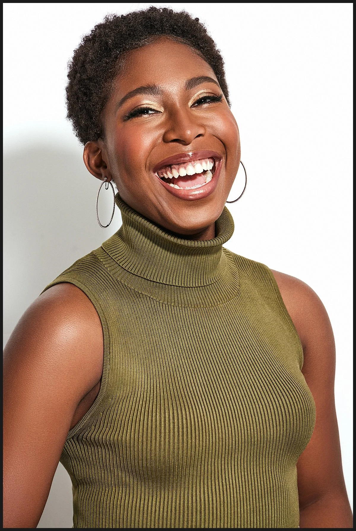 A smiling woman with short curly hair, wearing a green sleeveless turtleneck top and hoop earrings, posing against a white background.