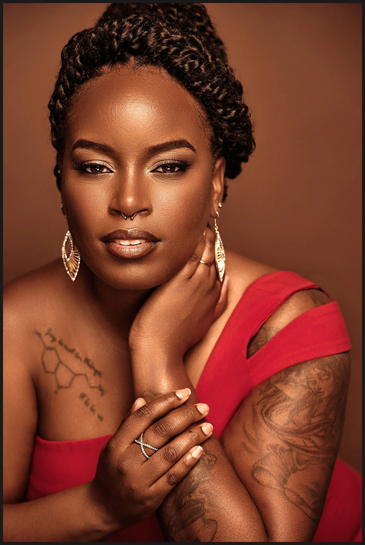 A portrait of a woman with dark skin, styled hair in twists, wearing a red off-shoulder top with cutout straps. She has gold jewelry, including earrings, a nose ring, and a ring on her finger. She has tattoos on her shoulder and arm and is gazing at 
