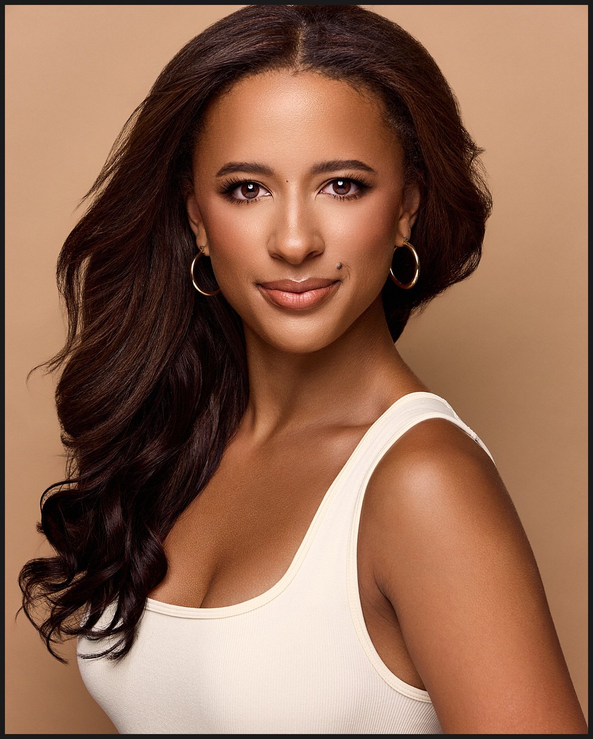 A woman with long, wavy dark hair wearing hoop earrings and a white tank top, looking at the camera with a neutral expression against a beige background.