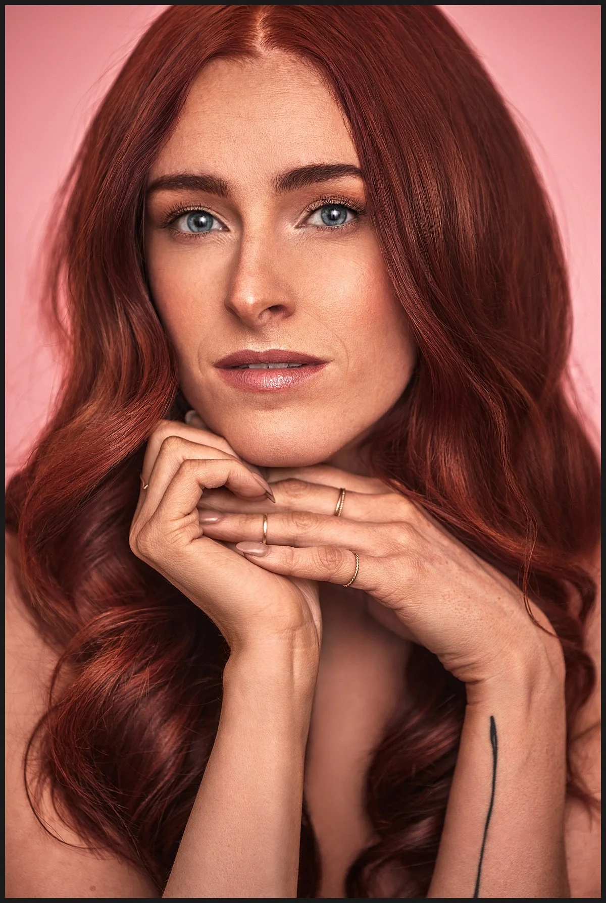 A close-up portrait of a woman with long, wavy red hair and blue eyes, resting her chin on her hands against a pink background.