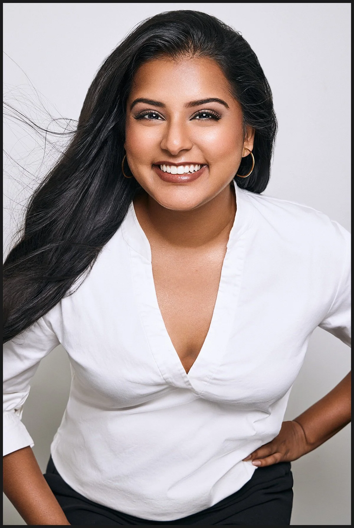 A woman with long black hair, wearing hoop earrings and a white blouse, smiling at the camera against a plain background.
