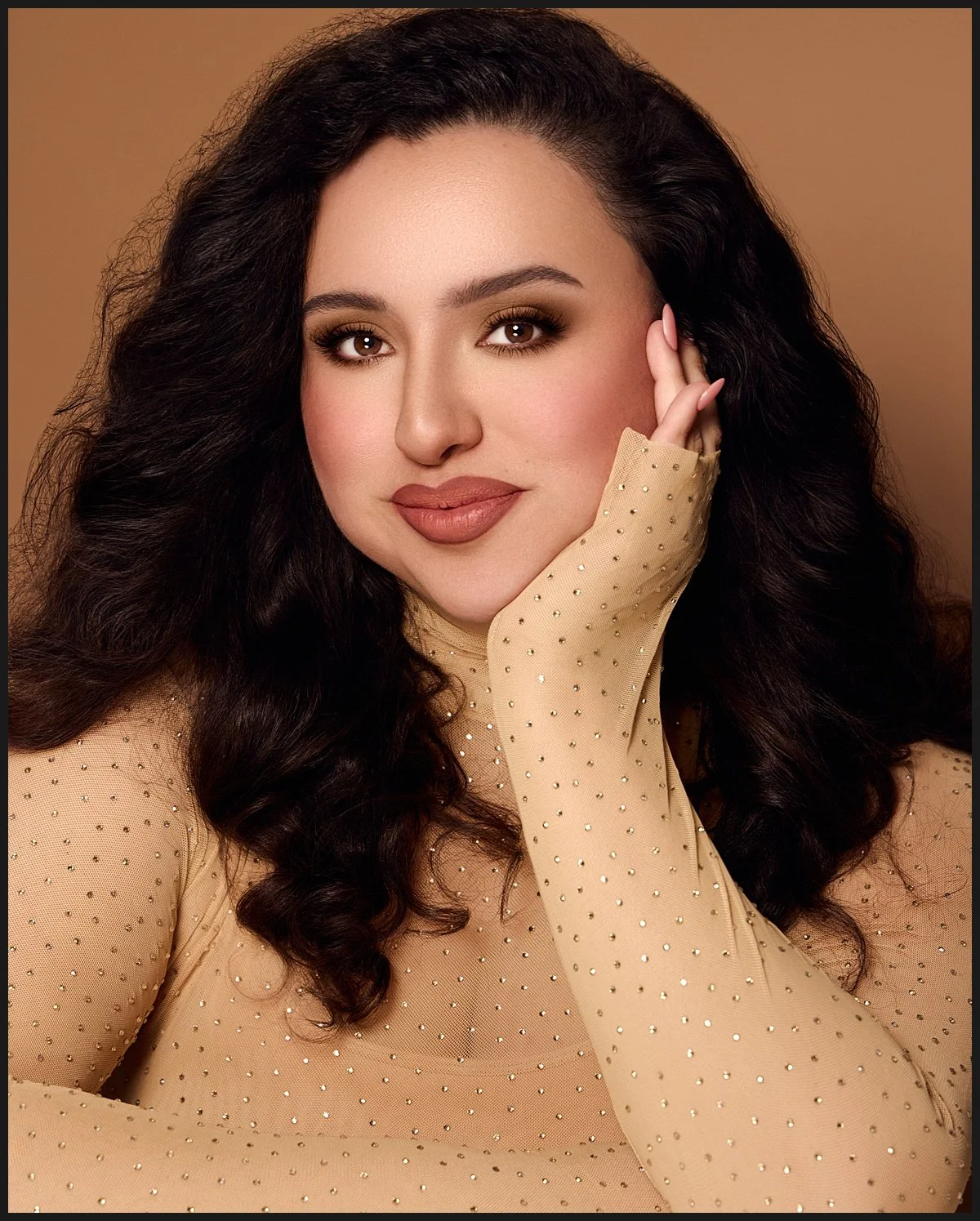 Close-up portrait of a woman with dark, curly hair, wearing a beige top with small, shiny embellishments, touching her face gently with her right hand.