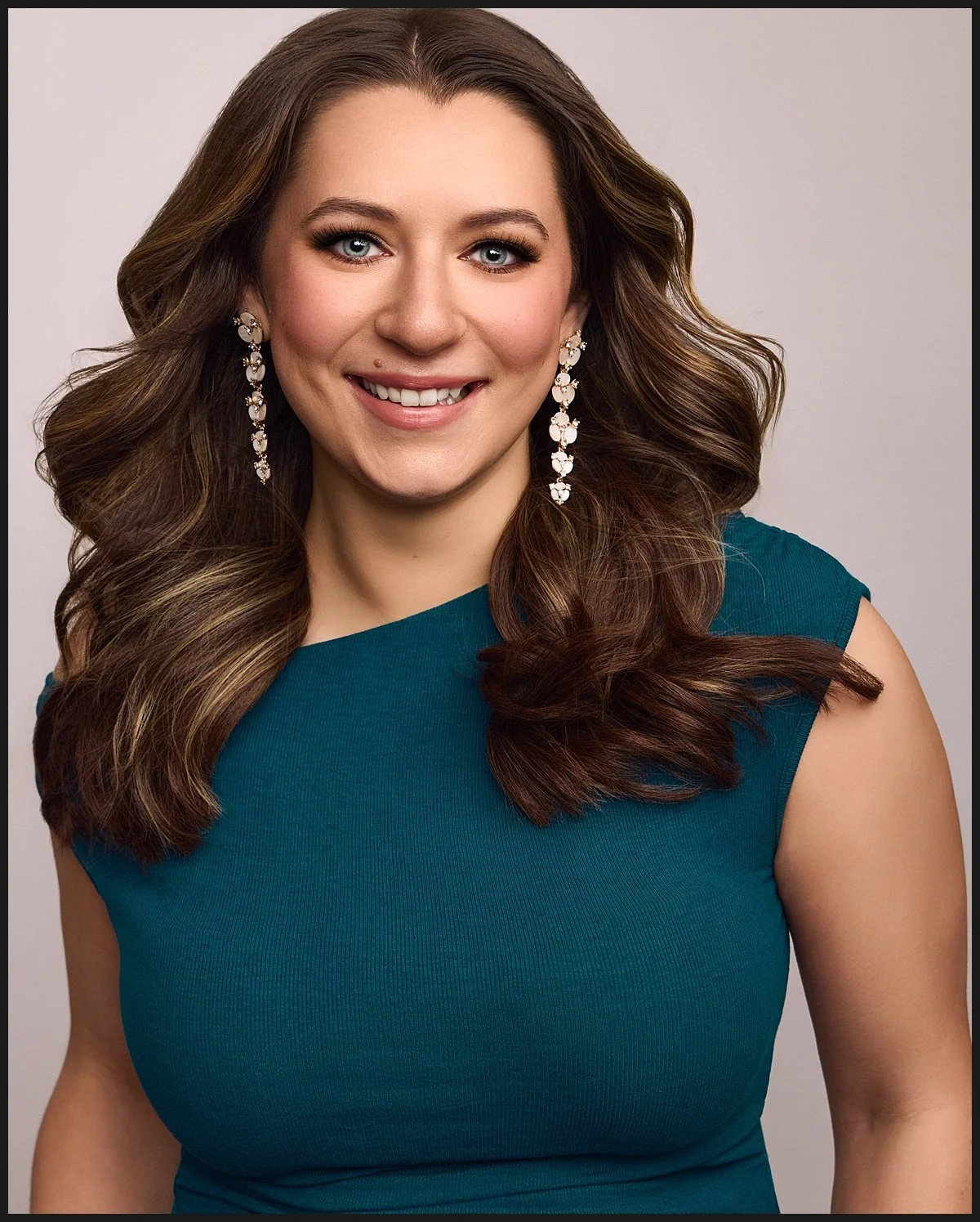 A woman with long, wavy brown hair wearing a teal sleeveless dress and long, dangling earrings, smiling at the camera against a plain light background.