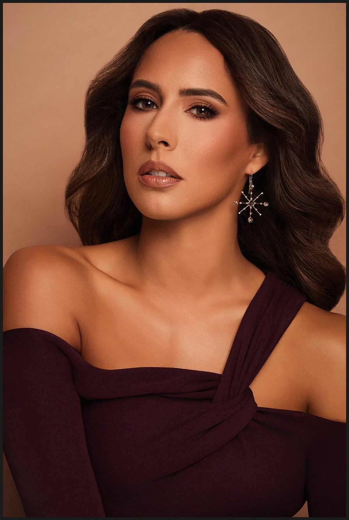 A woman with long wavy brown hair and light makeup, wearing a dark purple off-the-shoulder dress and a silver snowflake-shaped earring, posing against a beige background.