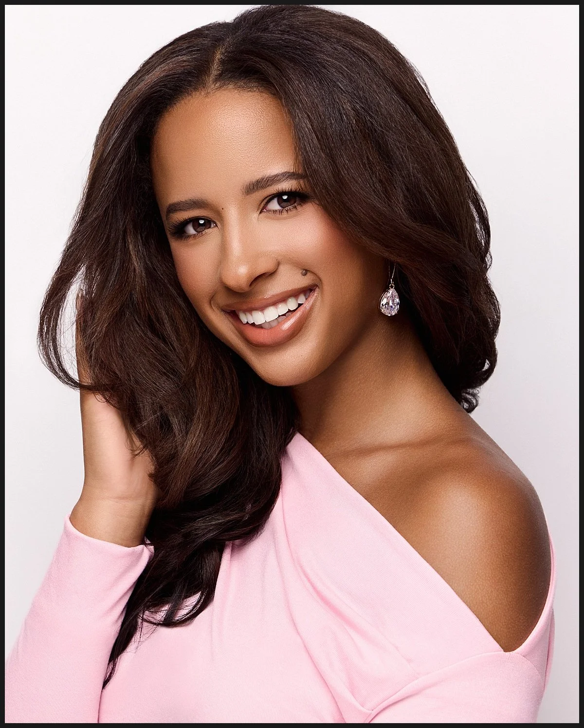 A woman with long wavy dark hair and medium skin tone, smiling, wearing a light pink off-the-shoulder top and teardrop earrings, with a white background.