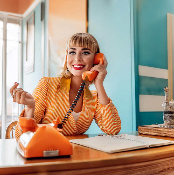 A woman smiling and talking on an orange rotary phone in a brightly colored room with blue and orange walls.
