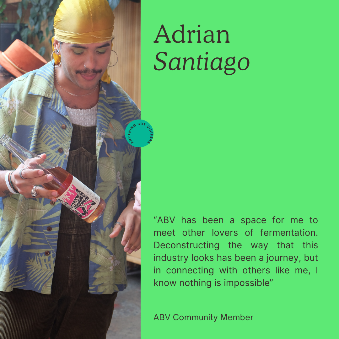 A person with a mustache and earrings wearing a yellow headscarf and colorful patterned shirt, holding a bottle of pink beverage, standing outdoors next to a wooden wall.