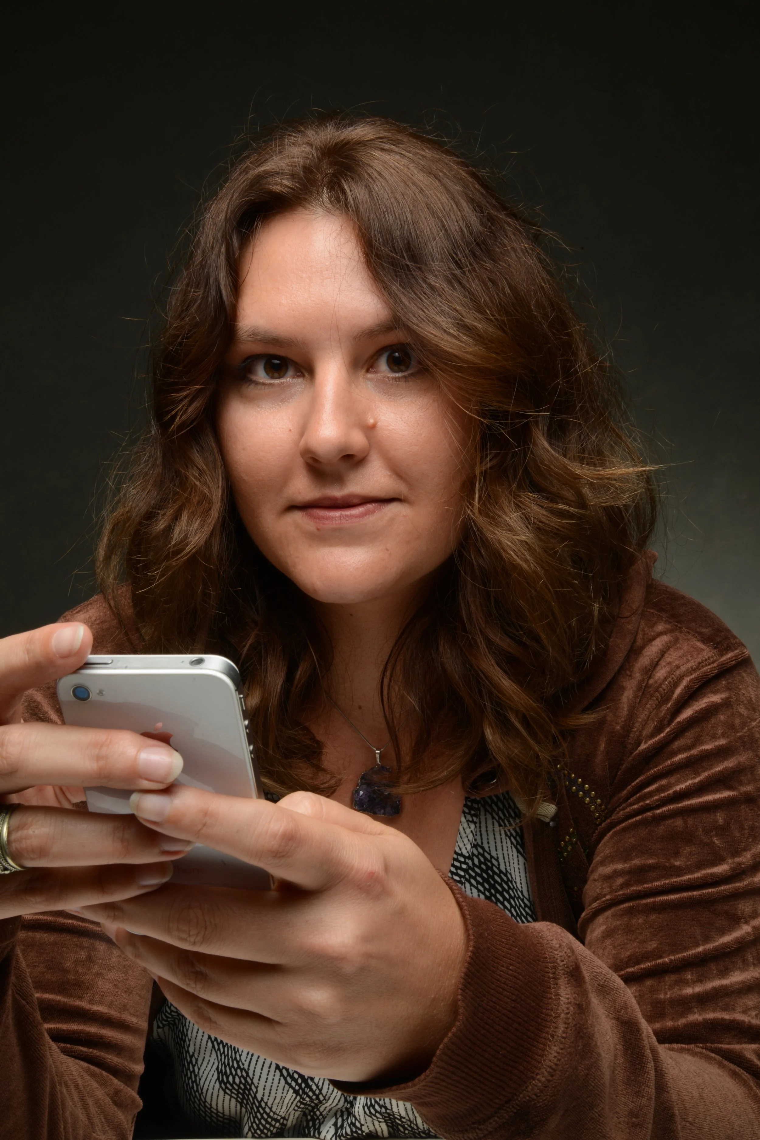 A woman with long, wavy brown hair is looking at her smartphone, wearing a brown jacket and a patterned top, against a dark background.