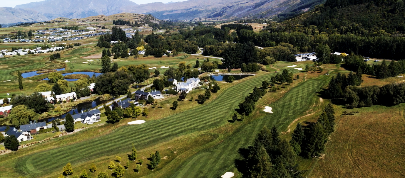 Aerial view of a golf course with sand traps, water hazards, and surrounding residential houses and trees, set against mountain backdrop.