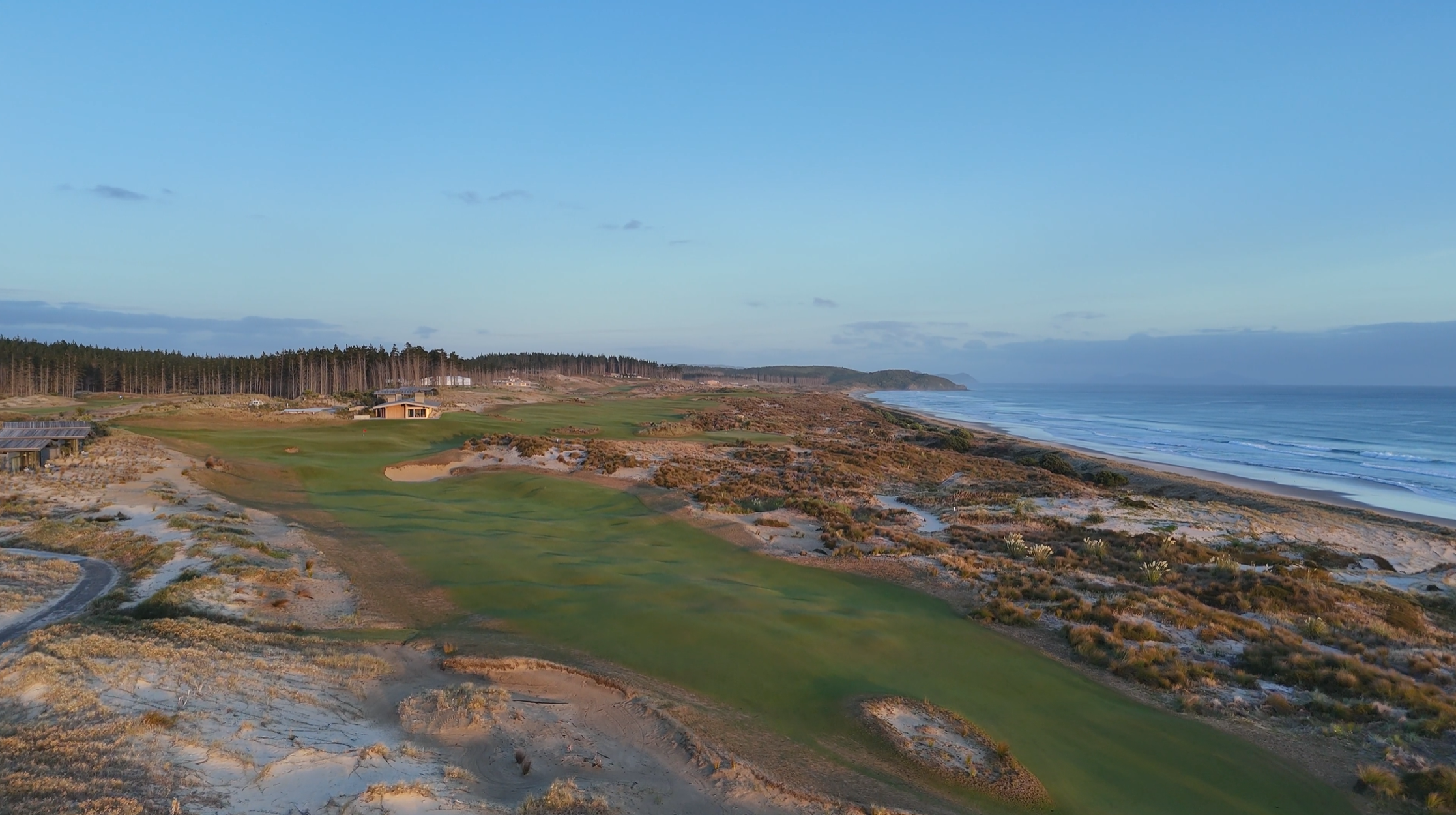 A coastal golf course with green fairways and sand dunes, overlooking the ocean with waves and a clear blue sky.