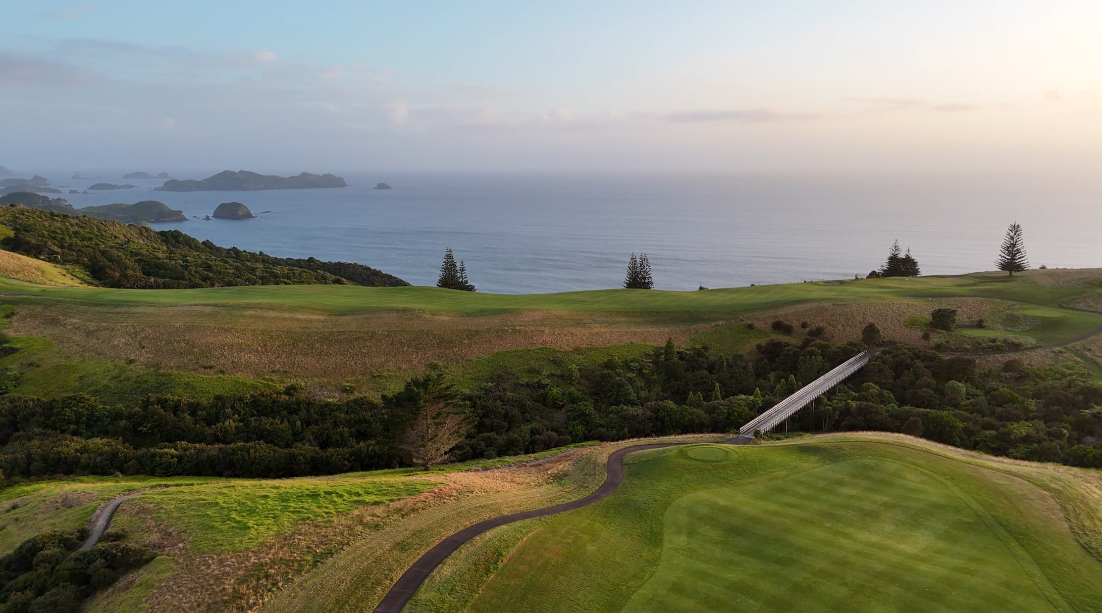 A scenic view of Kauri Cliffs golf course with green fairways, trees, and a bridge, overlooking the ocean with islands in the distance during sunset.