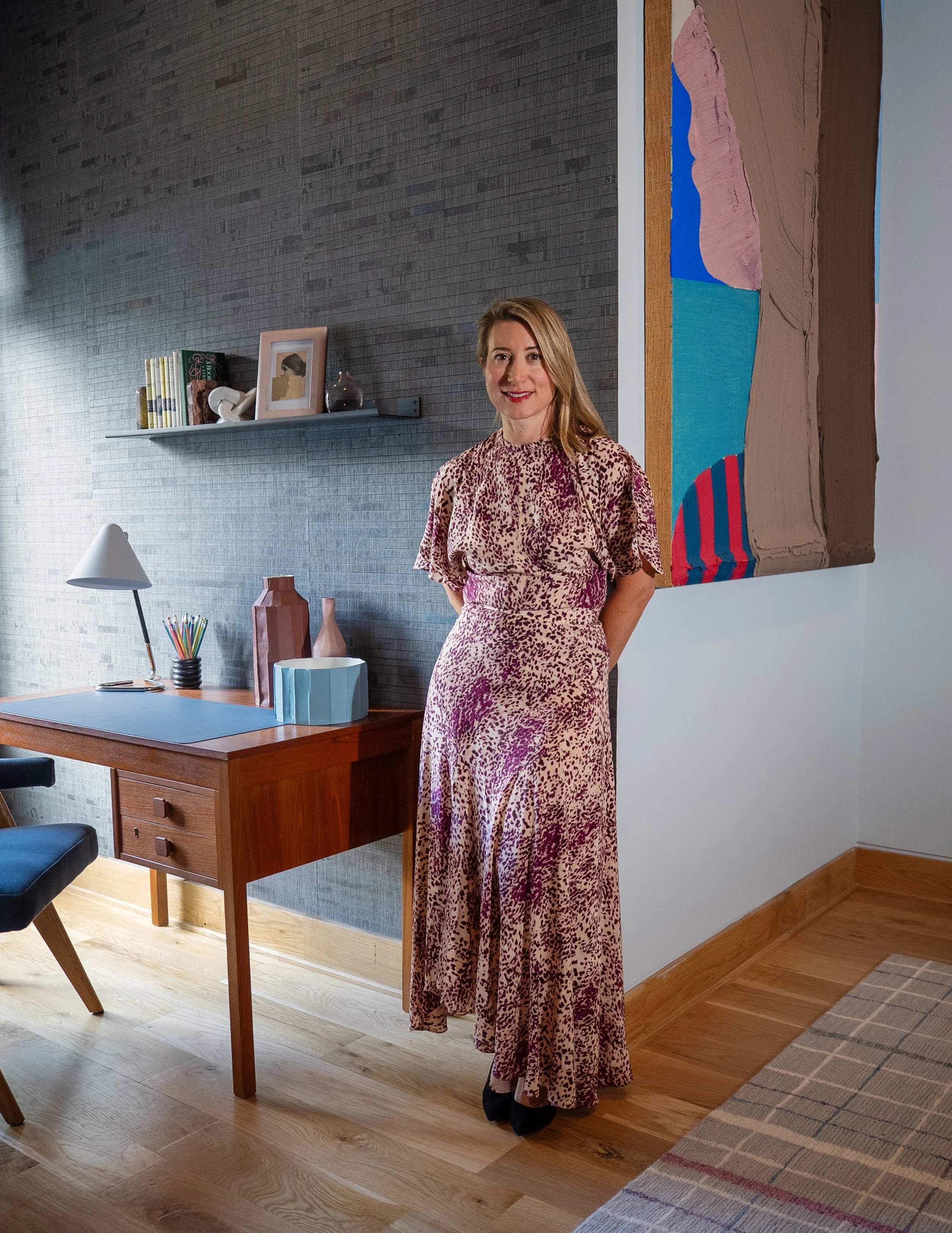 A woman in a pink and purple animal print dress stands in a modern home office, smiling. There is a wooden desk with a desk lamp, vases, and pencils, and a gray textured wall with a floating shelf and abstract artwork.