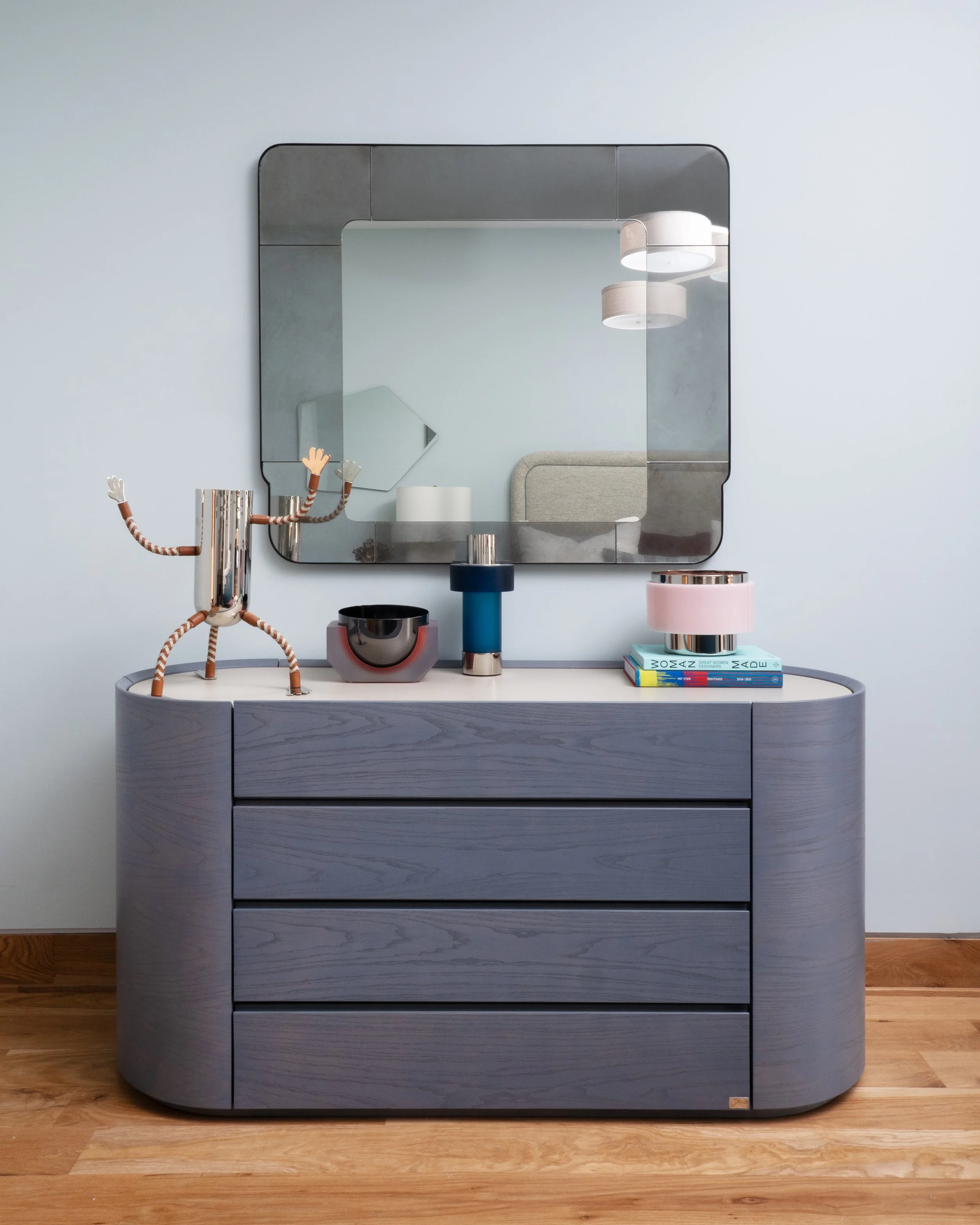 A modern dresser with a mirror above it, decorated with contemporary accessories including a whimsical robot sculpture, various small lamps, and a stack of colorful books, set against a light blue wall and wooden floor.