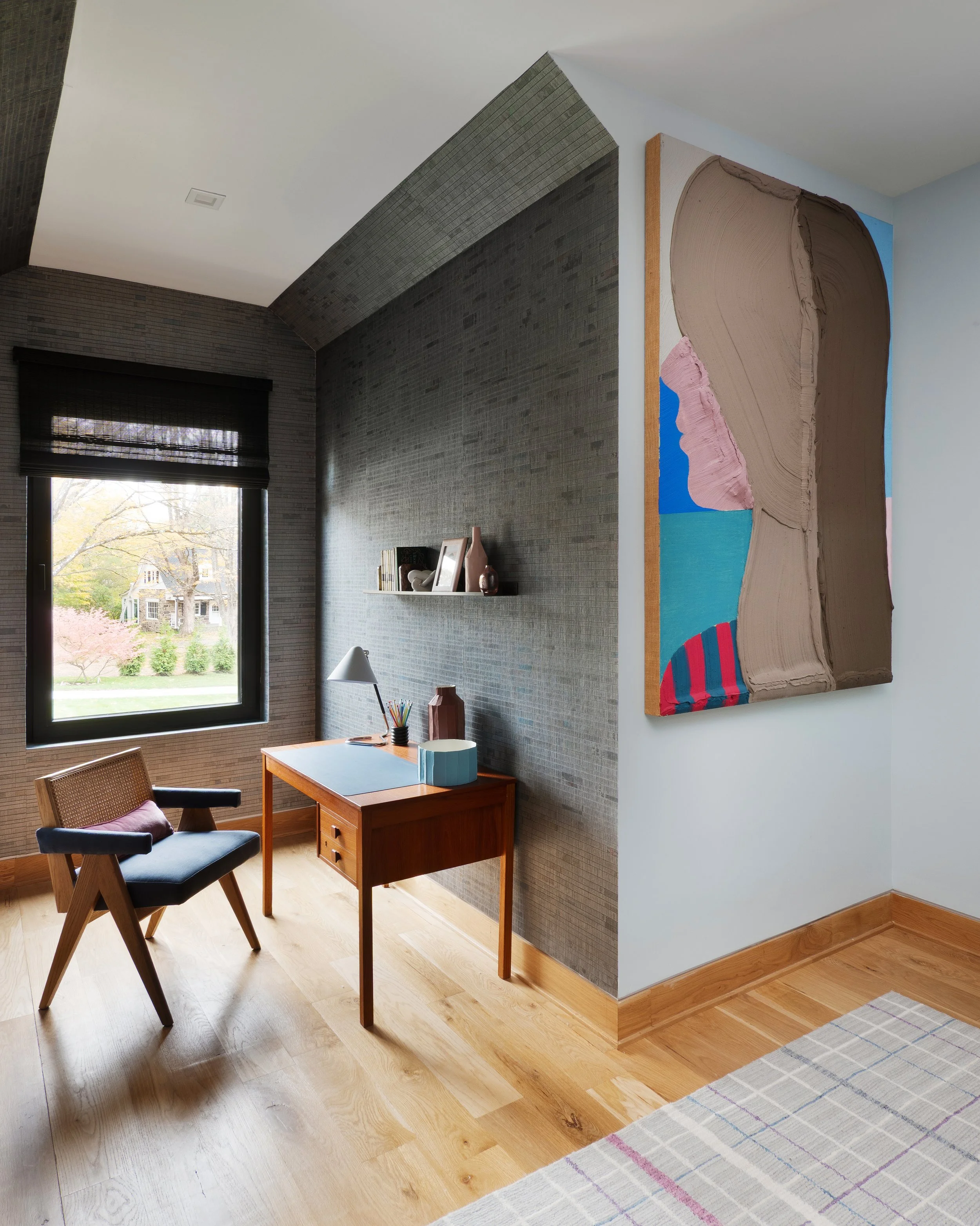 A home office with a wooden desk, a black and wood chair, a window with a black shade, and a colorful abstract painting on the white wall.