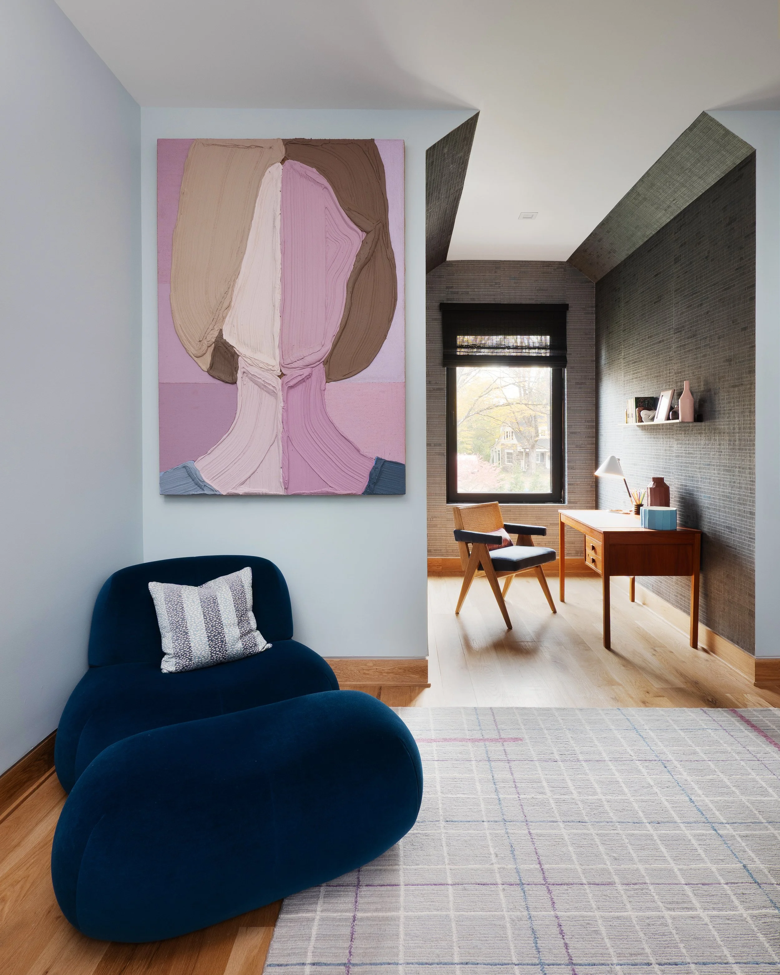 Modern living room with a large abstract portrait painting on the wall, a blue velvet armchair with a patterned pillow, a wooden desk with a chair near a window, and a light-colored rug on hardwood floor.