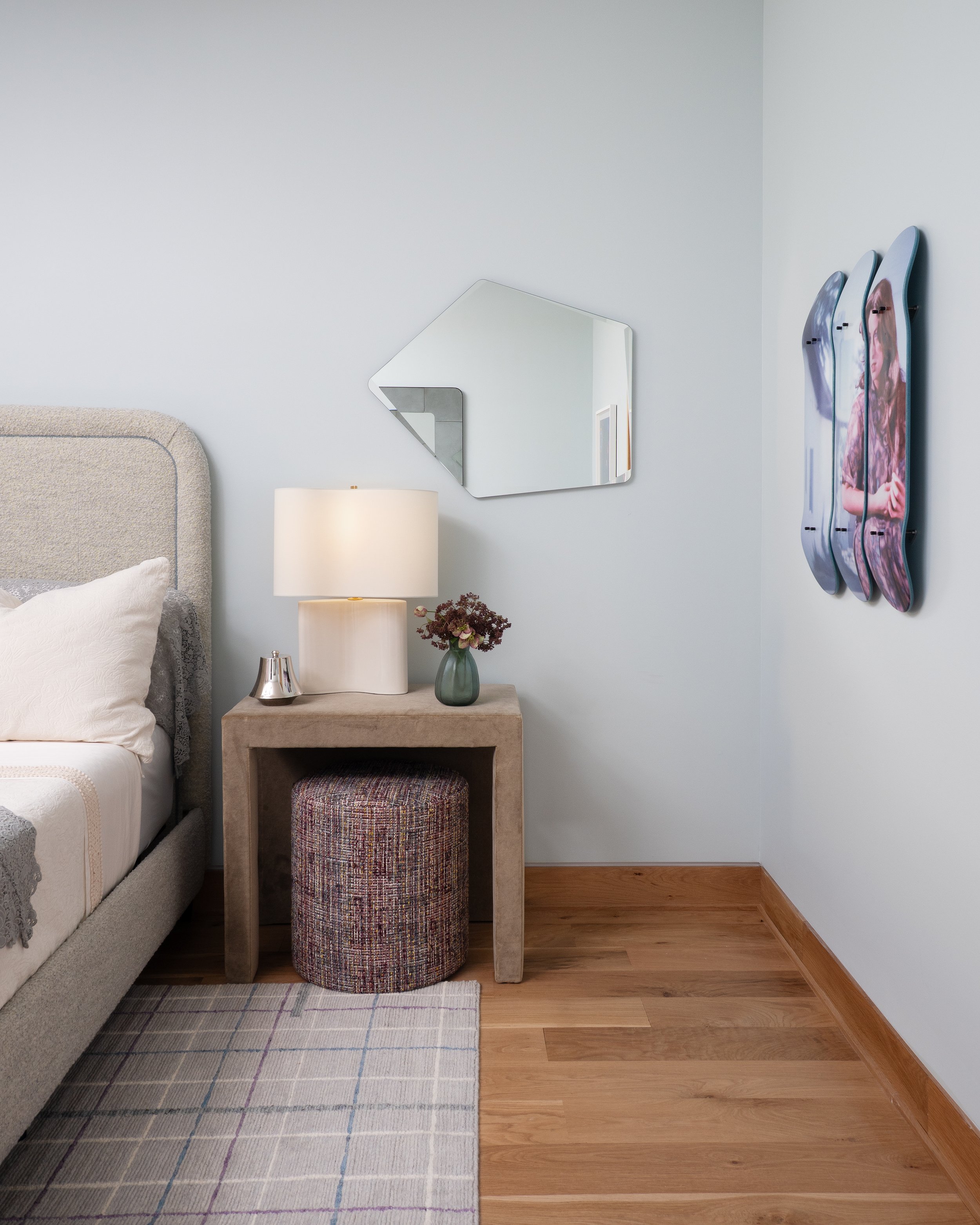 A bedroom corner with a gray upholstered bed, a beige bedside table with a lamp, a small flower vase, a mirror on the wall, and skateboards hanging on the wall.