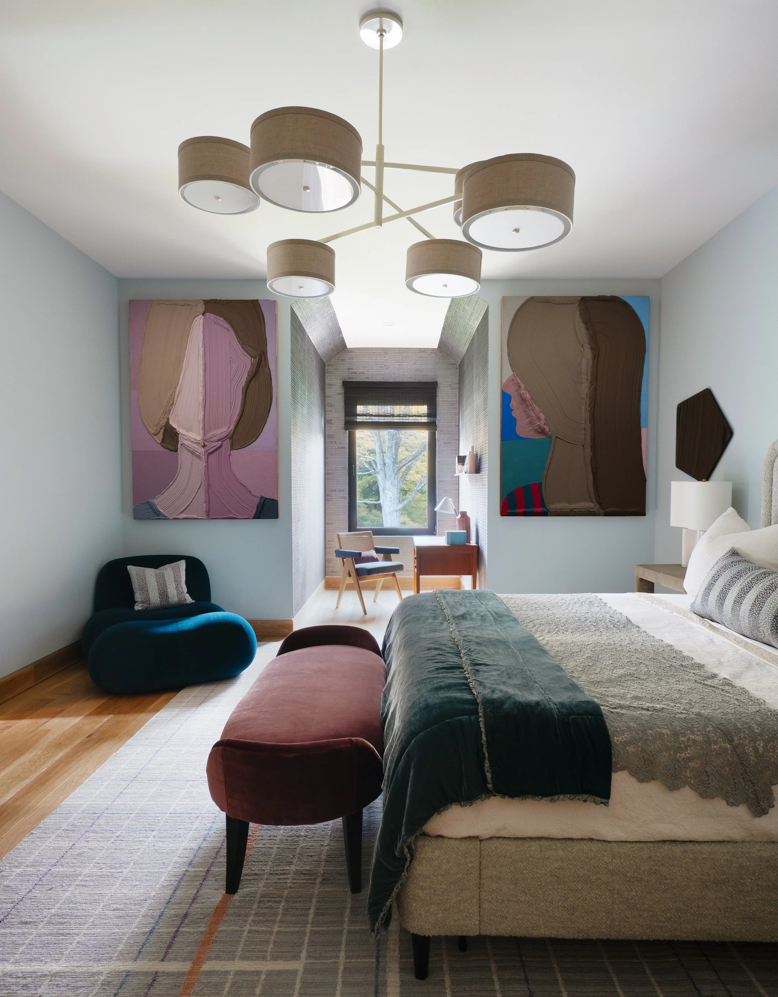 Modern bedroom with a bed, velvet bench, and two colorful abstract portraits on the wall, with a sitting area by the window