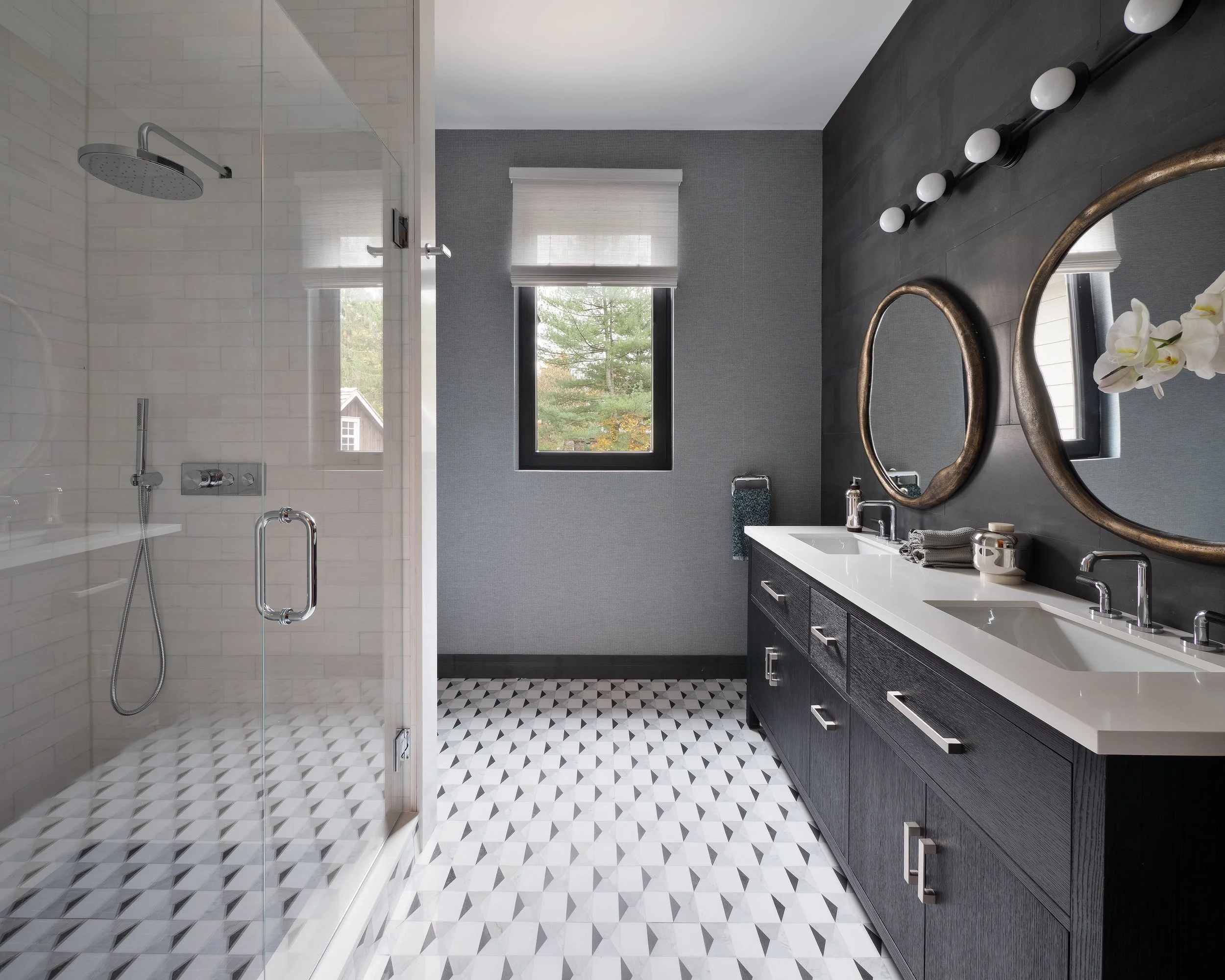 Modern bathroom with a glass-enclosed shower on the left, featuring a rainfall showerhead, and a dark wooden double vanity with two sinks on the right. The vanity has two oval mirrors, and a black wall with a row of globe lights. Two windows provide natural light, one with a white blind and the other with a view of trees outside. The floor has a geometric black and white tile pattern.