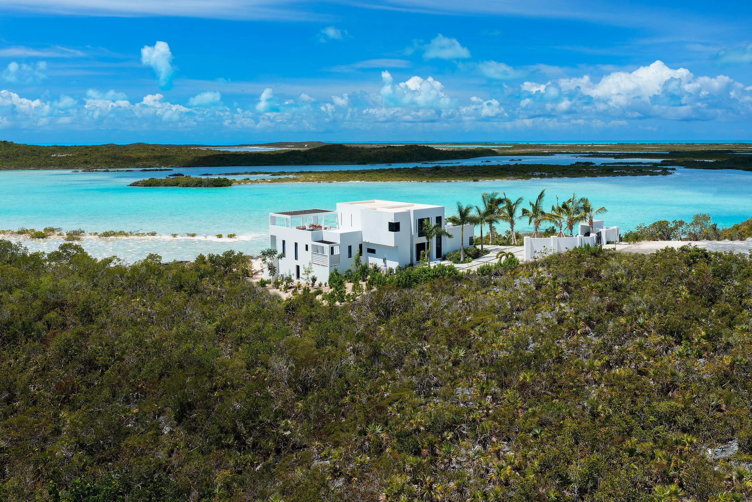 A modern white house with flat roofs and a rooftop terrace situated on a sandy riverbank with tropical trees and bushes, overlooking a turquoise ocean and distant islands under a partly cloudy blue sky.