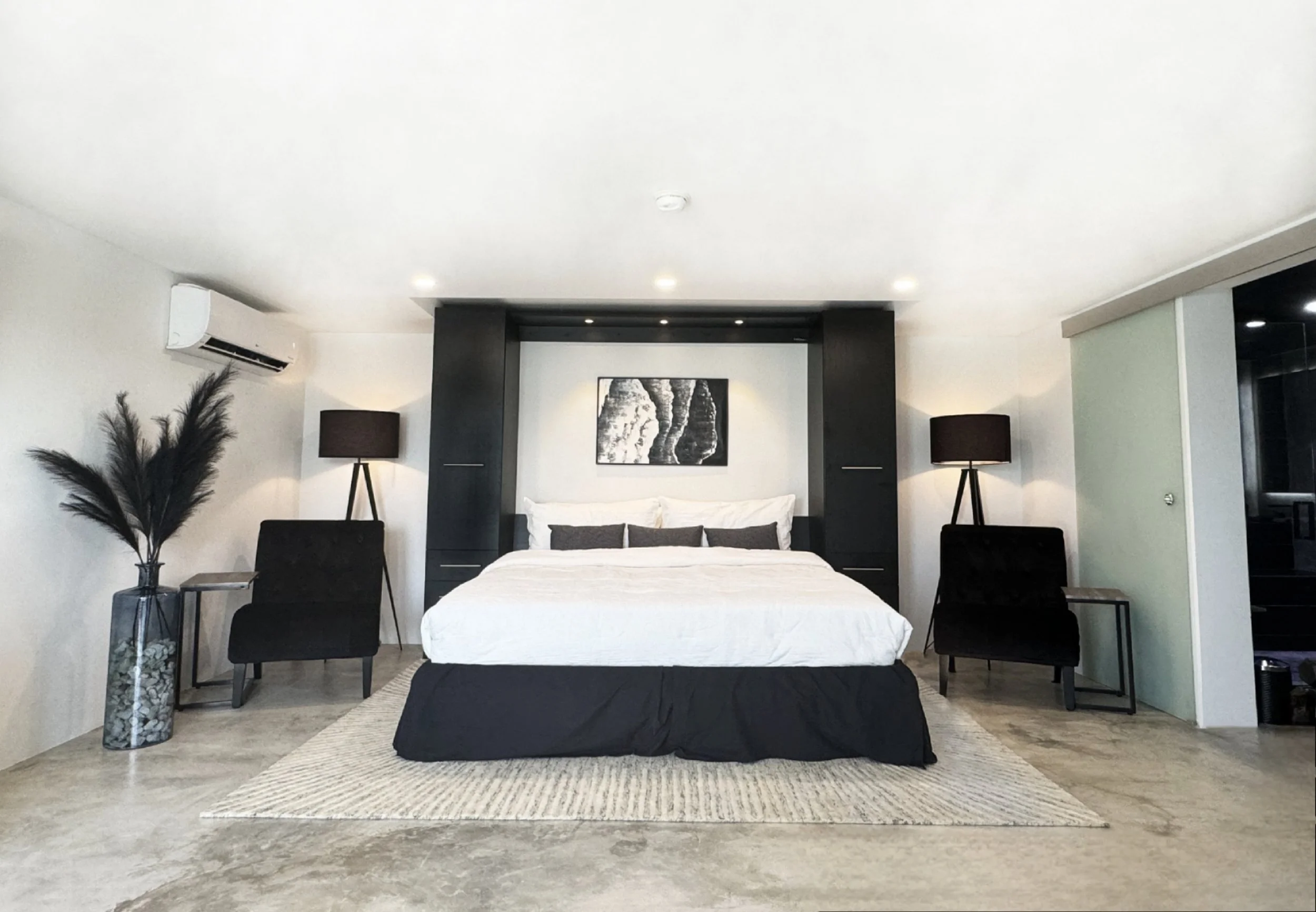 Modern bedroom with white walls, black accents, a large bed with white bedding and black pillows, black armchairs, and black lamps on side tables, decor includes a black and white artwork above the bed and a tall vase with black pampas grass.